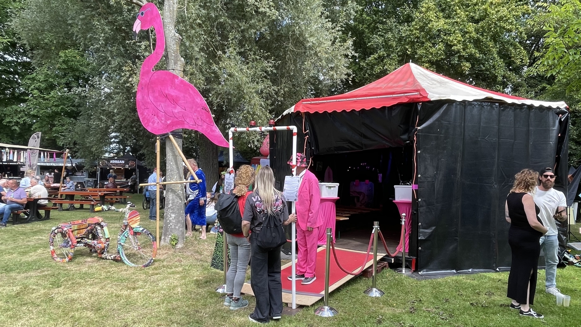 Een tent met een rood-wit dak en een groot roze flamingo-teken ervoor, omringd door mensen op een grasveld tijdens een evenement. Ernaast staan een kleurrijk versierde fiets en een man in een felroze pak. Op de achtergrond zitten mensen op picknickbanken.