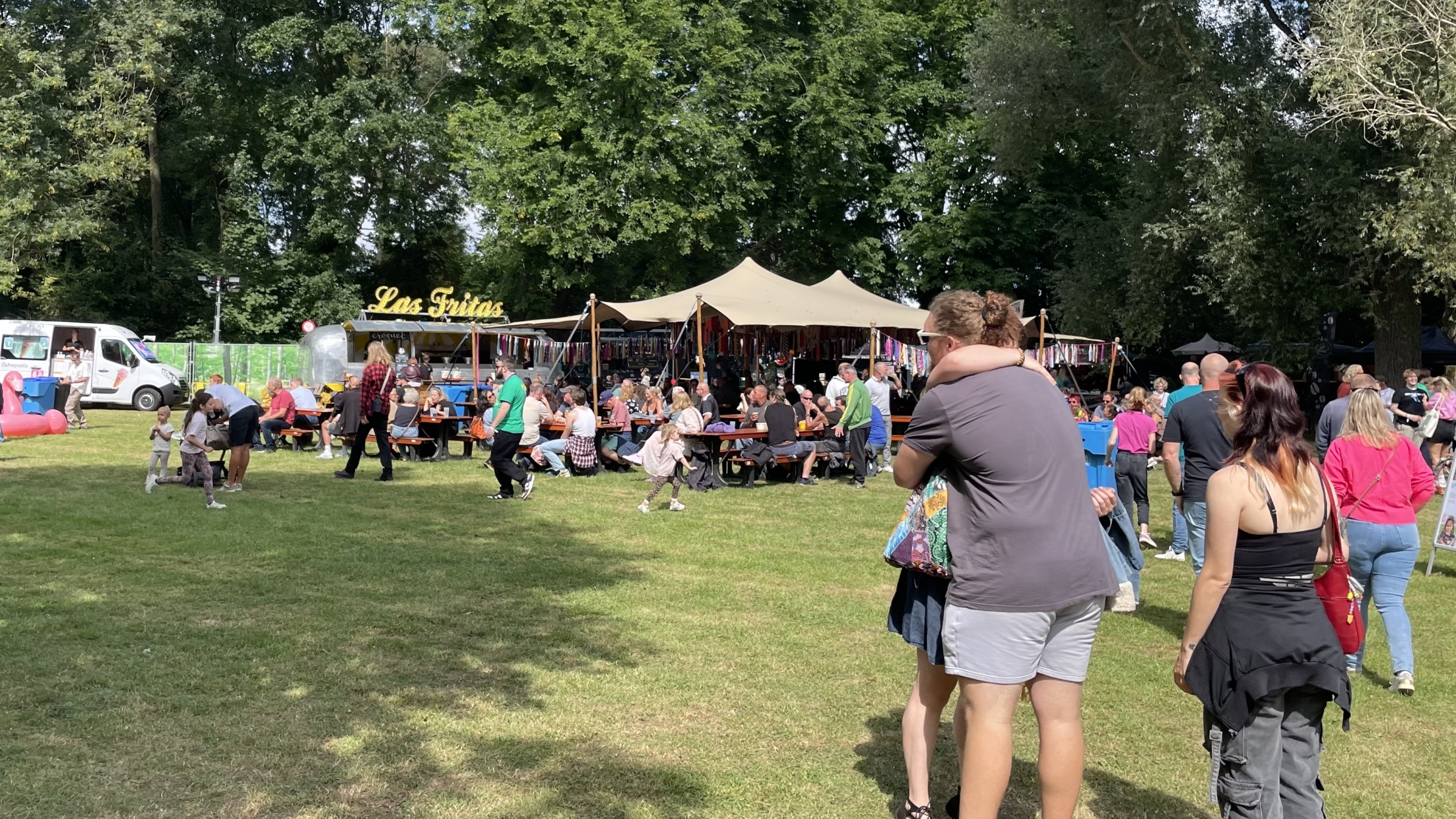 Mensen genieten van een buitenfestival met eetkraampjes, tafels en stoelen onder een tent, omgeven door bomen.