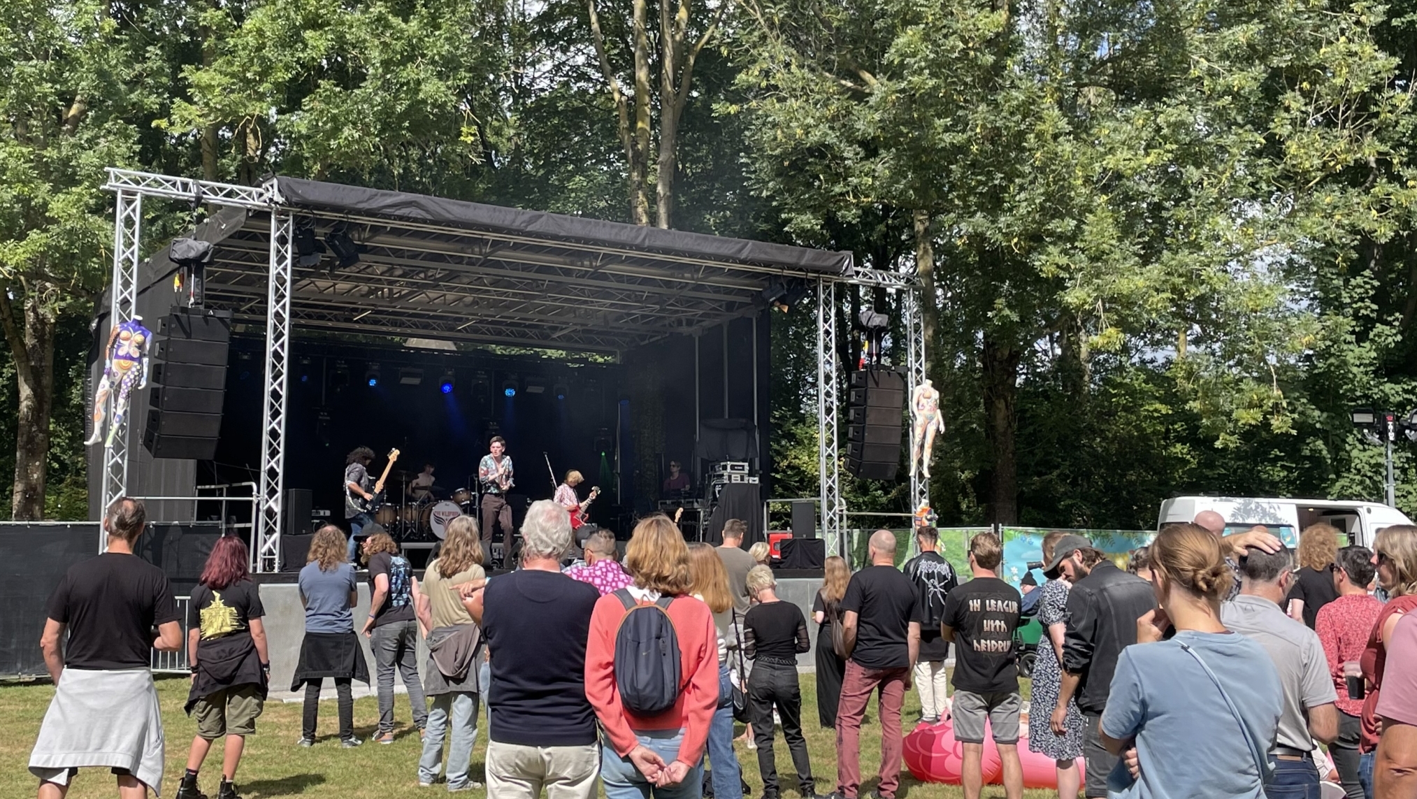 Mensen voor een buitenpodium kijken naar een optreden van een band, omringd door bomen en een zonnige lucht.