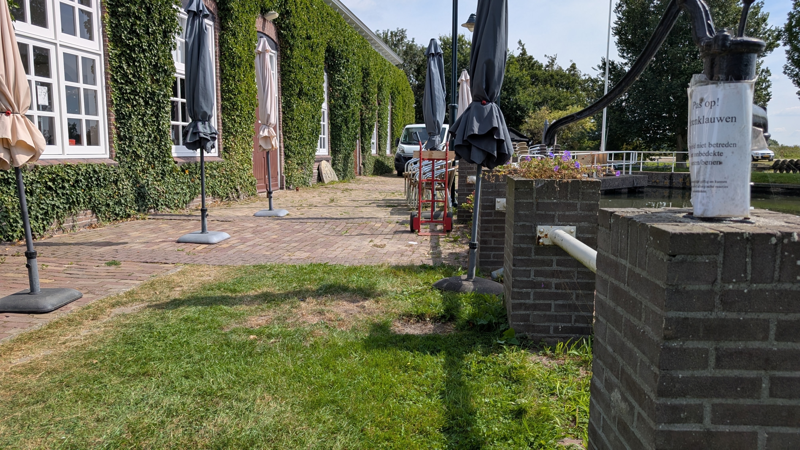 Terras met opgevouwen parasols naast een gebouw bedekt met klimop, en een bordje met waarschuwing voor kattenklauwen aan een muur; op de achtergrond is een geparkeerde busje te zien.