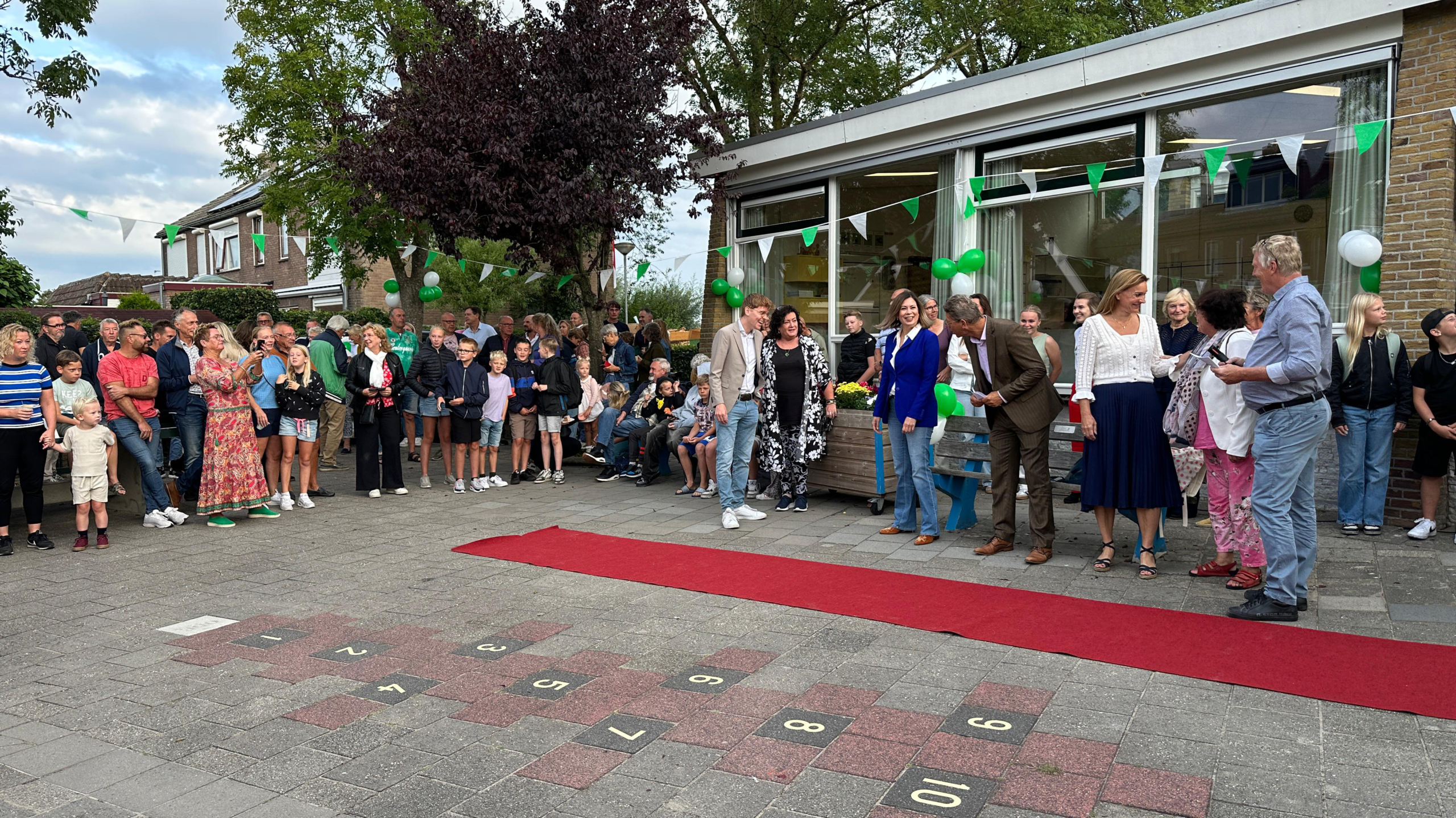 Een groep mensen staat buiten bij een feestelijke gelegenheid met groene decoraties en een rode loper op de grond. Sommigen maken foto's, terwijl anderen kijken of praten. Er zijn volwassenen en kinderen aanwezig bij het evenement.