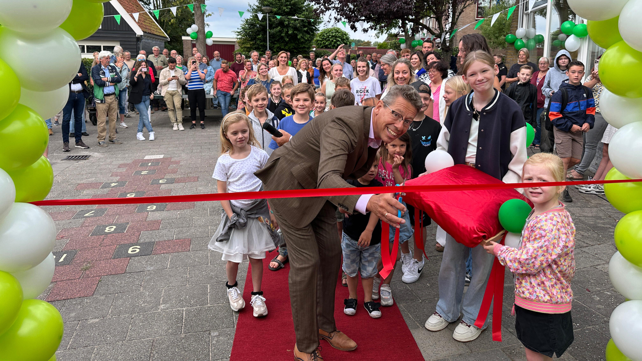Een man in een pak knipt een rode lint door bij een openingsceremonie, omringd door blije kinderen en toeschouwers. De achtergrond is versierd met groene en witte ballonnen en vlaggetjes.