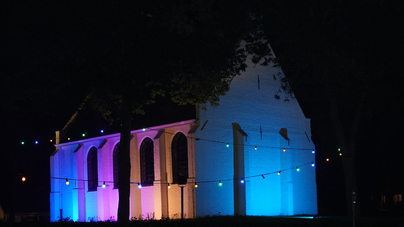 Verlichte kerk in de nacht met gekleurde verlichting; de muren zijn verlicht in blauw en paars, omlijst door kleurrijke prikkabelverlichting.