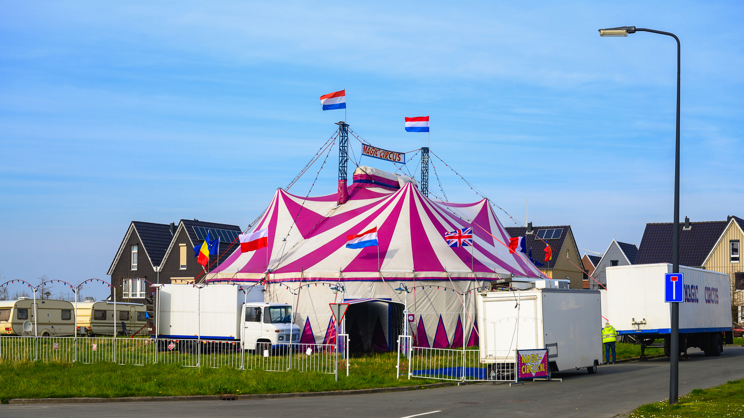 Roze-witte circustent met vlaggen van Nederland en het Verenigd Koninkrijk, omgeven door woonhuizen en geparkeerde caravans.