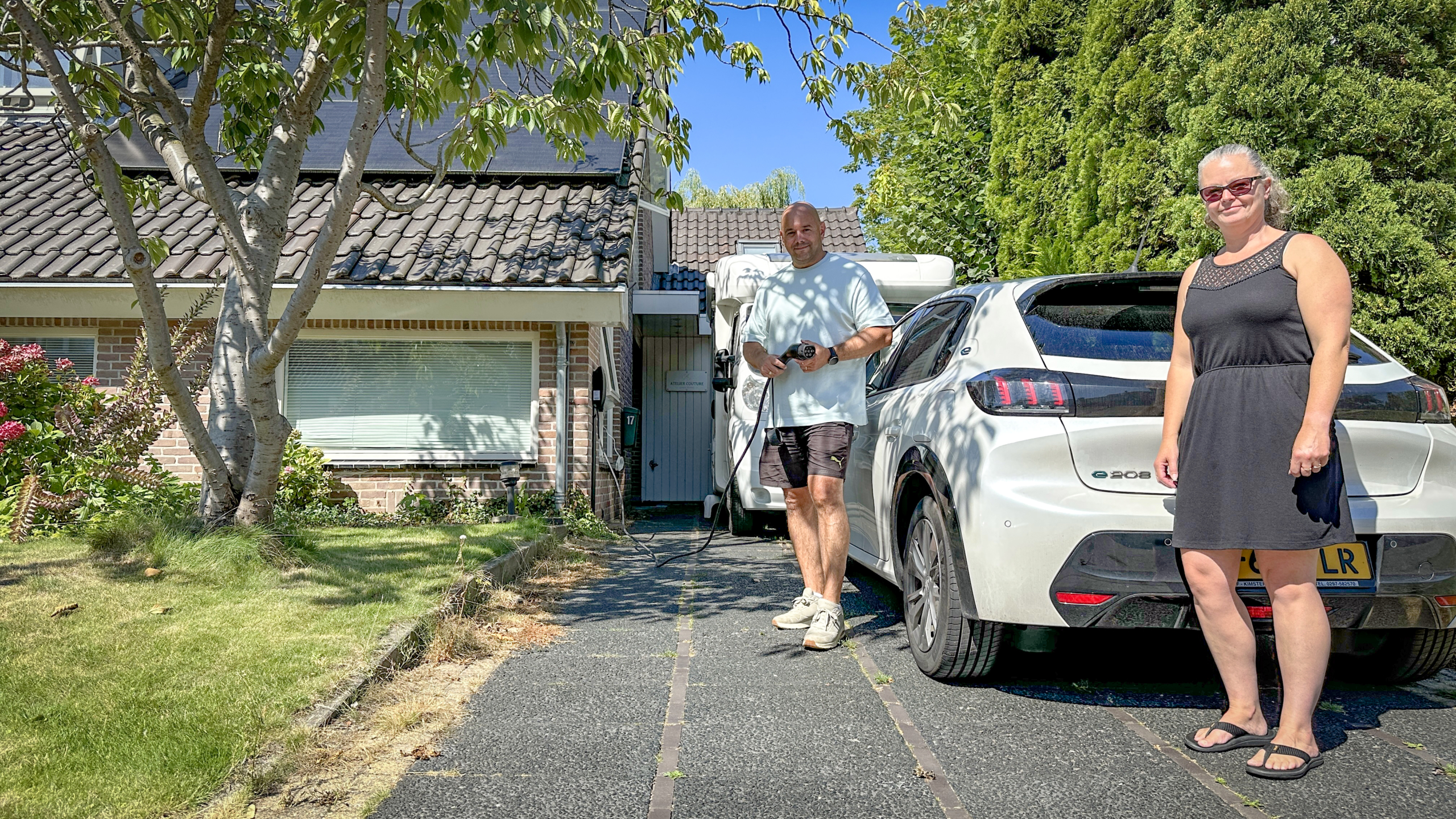 Een man en een vrouw staan buiten naast een witte elektrische auto die geparkeerd staat op een oprit voor een huis met zonnepanelen op het dak en een camper op de achtergrond. De man houdt een oplaadkabel vast, terwijl de vrouw glimlacht naar de camera. Er is gras en een boom in de voortuin zichtbaar.