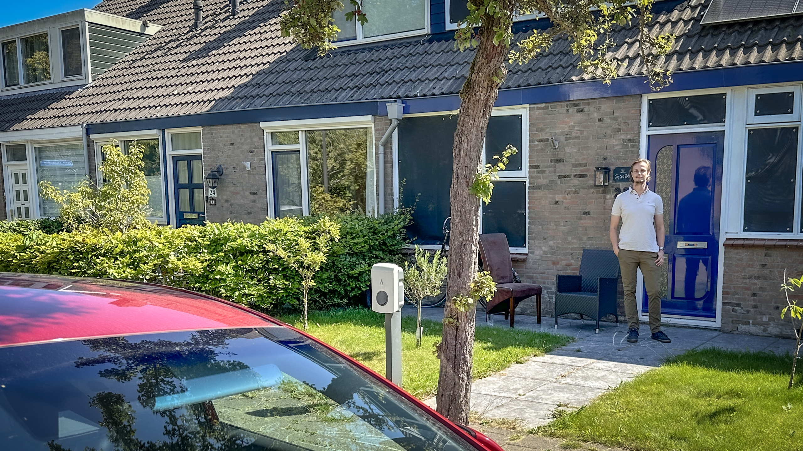 Een man staat voor een huis met een blauwe deur, omgeven door een heg en een kleine tuin, met een elektrische laadpaal en een deel van een rode auto op de voorgrond.