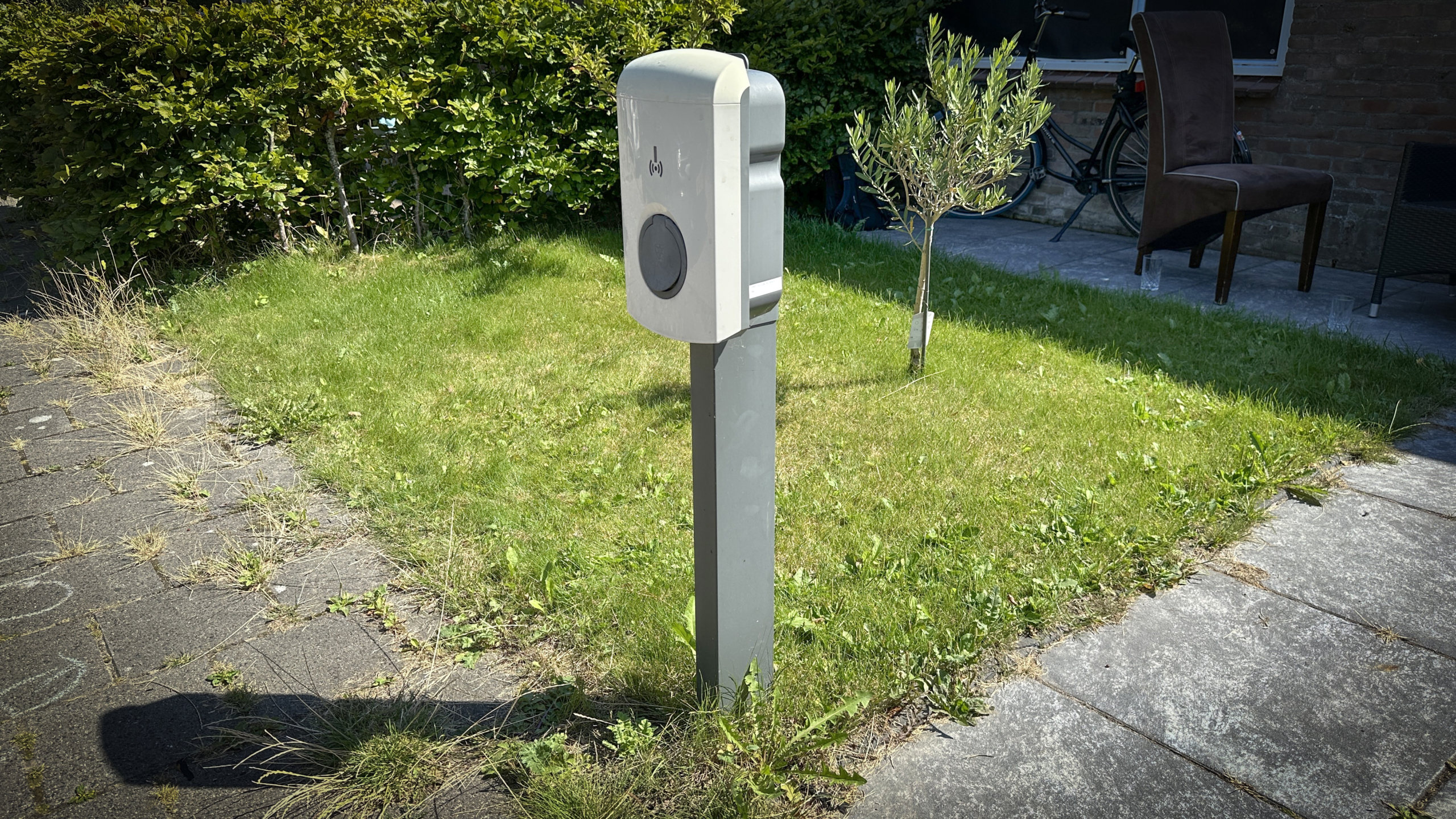Oplaadpaal voor elektrische auto's op een grasveld naast een geplaveid pad en een kleine boom, met een fiets en stoel in de achtergrond.