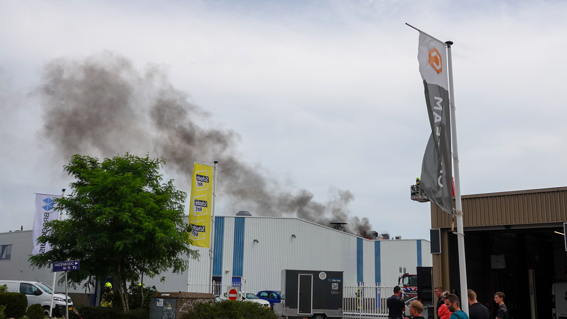 Een fabriek met rook die opstijgt uit het gebouw, terwijl brandweerlieden in actie zijn. In de voorgrond staan enkele mensen en zijn er vlaggen te zien.