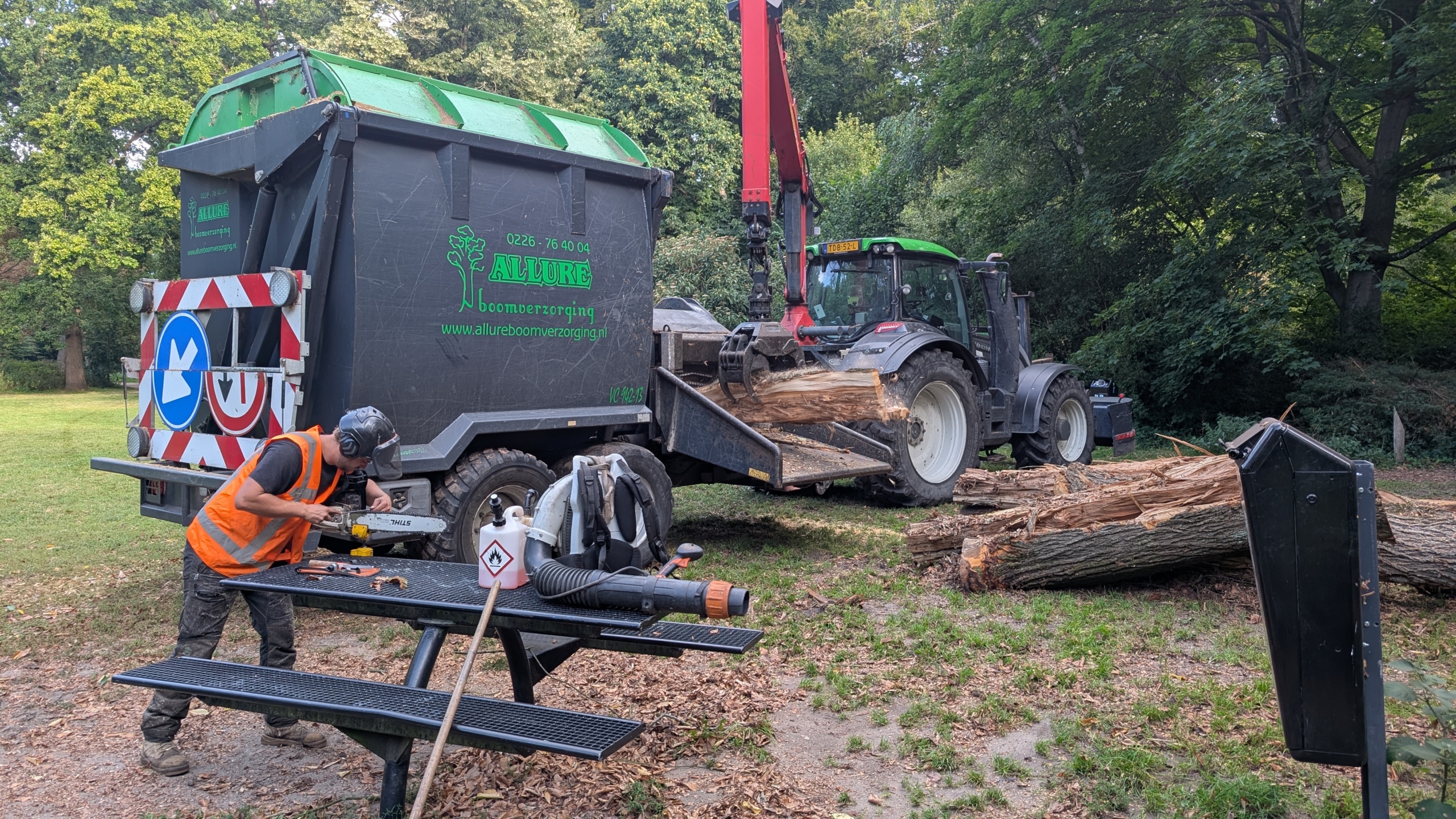 Een man in een oranje veiligheidshesje werkt met een kettingzaag naast een vrachtwagen met boomstammen, terwijl een tractor boomstammen laadt in een groene omgeving.