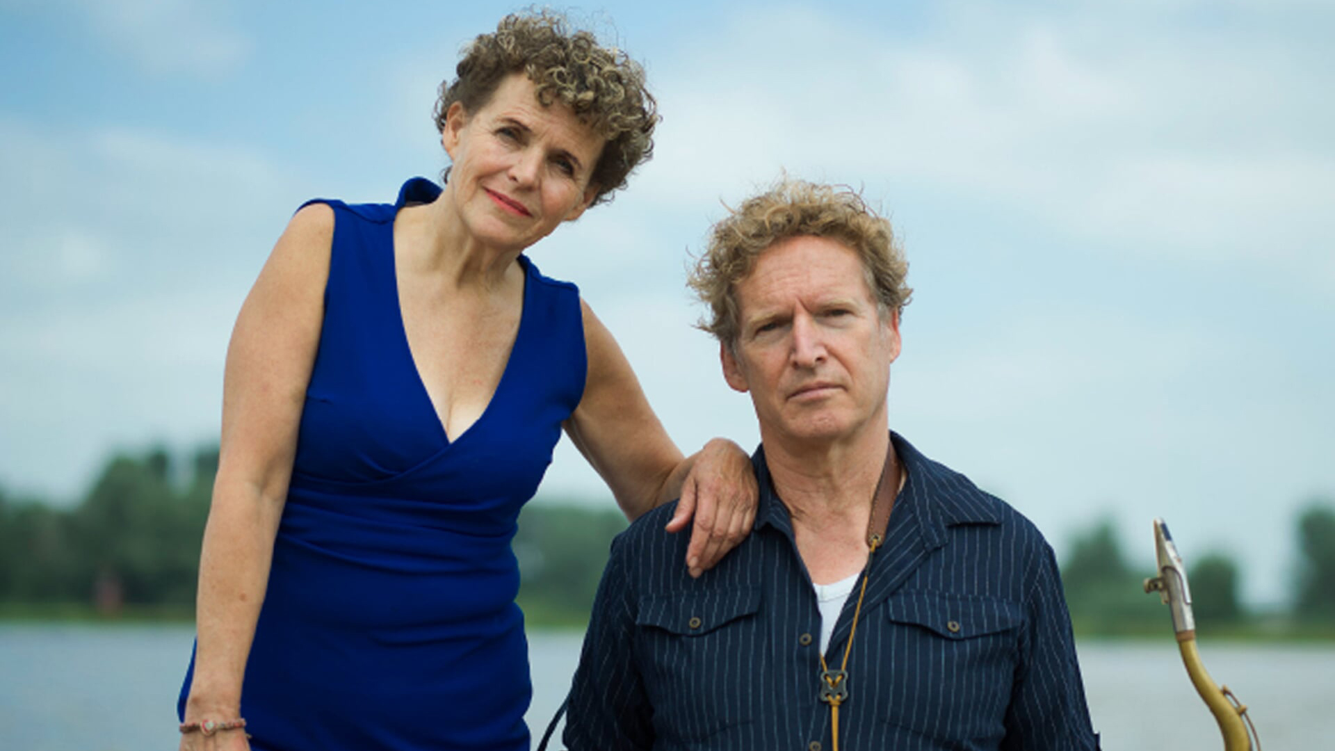 Een vrouw in een blauwe jurk staat naast een man met een gestreept overhemd, buiten met een wazige achtergrond van bomen en water.