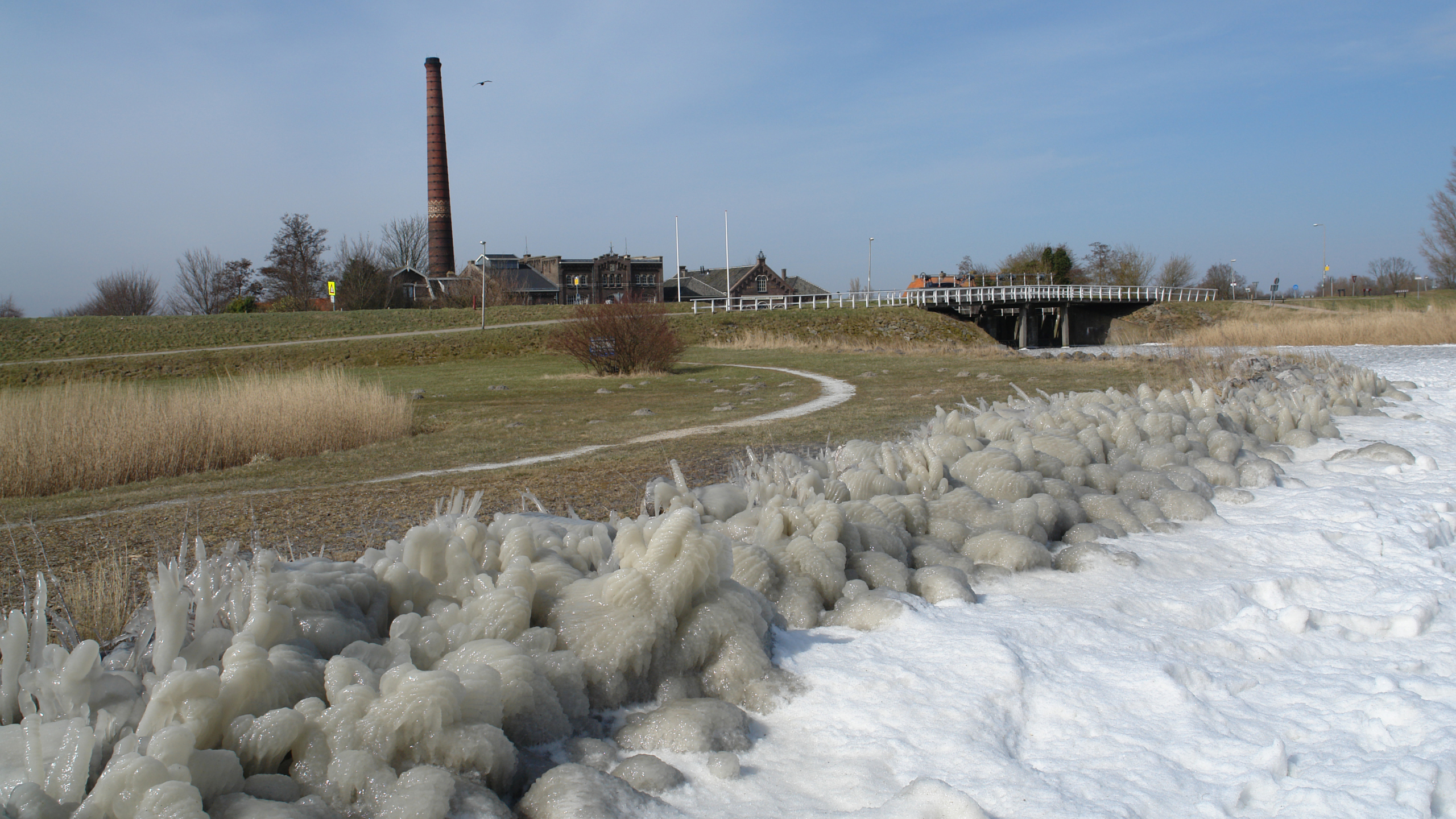 Een bevroren landschap met ijsformaties op de voorgrond en een industriële schoorsteen op de achtergrond naast een brug en een aantal gebouwen.