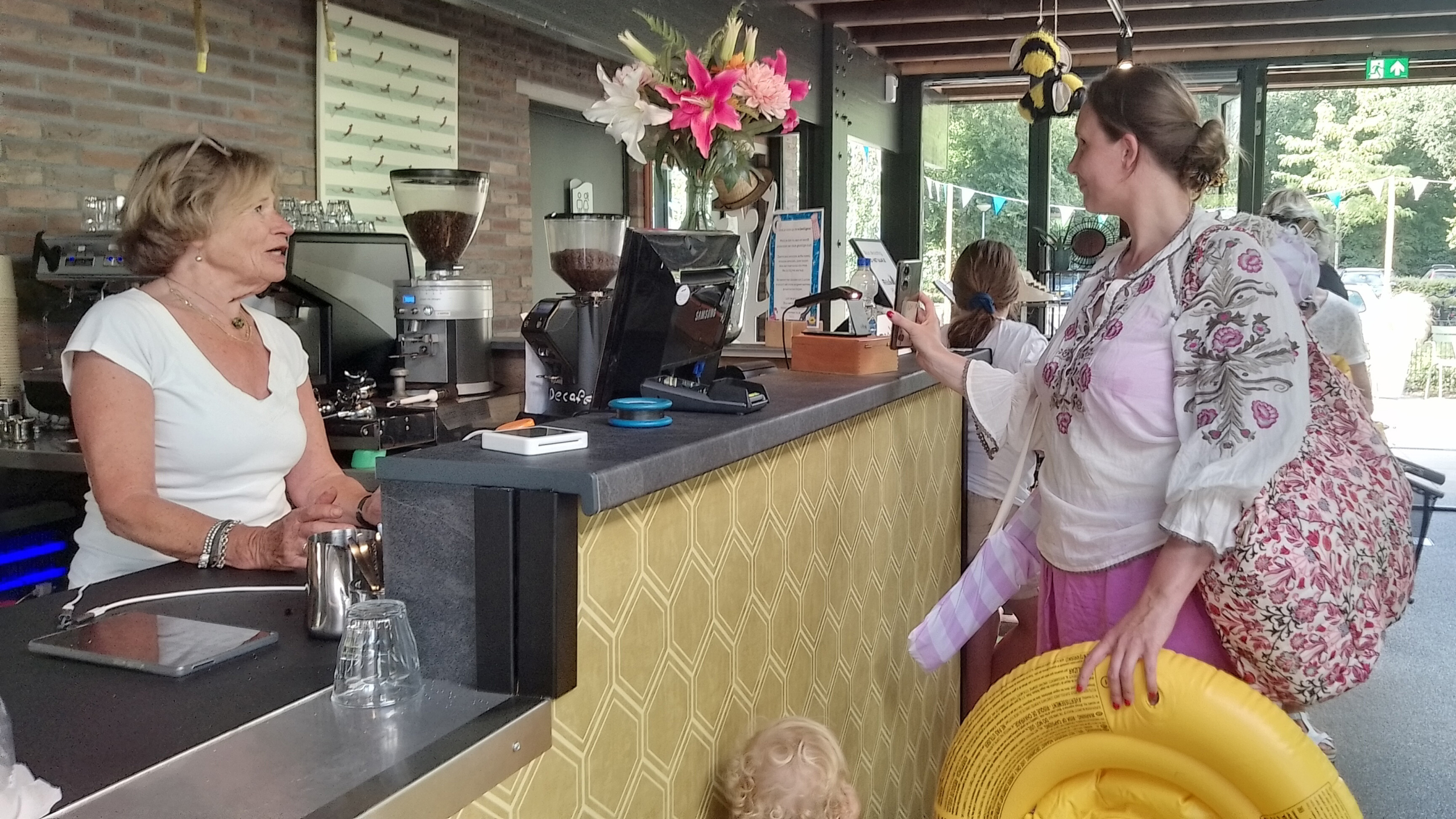 Een vrouw in een gebloemde blouse spreekt met een vrouw achter een cafételler, terwijl ze een opblaasbare zwemband vasthoudt en verschillende spullen bevat in een tas. De teller heeft een koffiezetapparaat en een boeket bloemen.