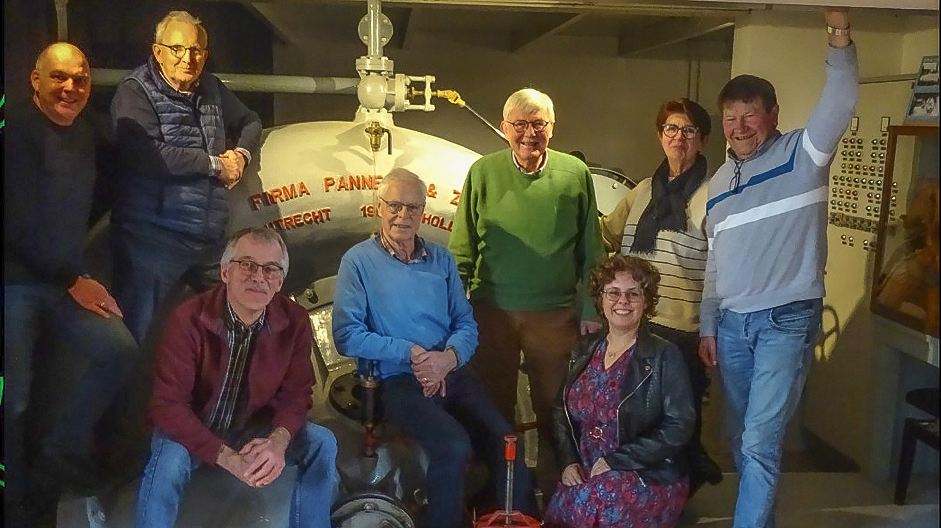 Groep volwassenen poseert voor een ouderwetse machine met opschrift "Firma Pannevis & Zoon - Utrecht 1911 Holland".