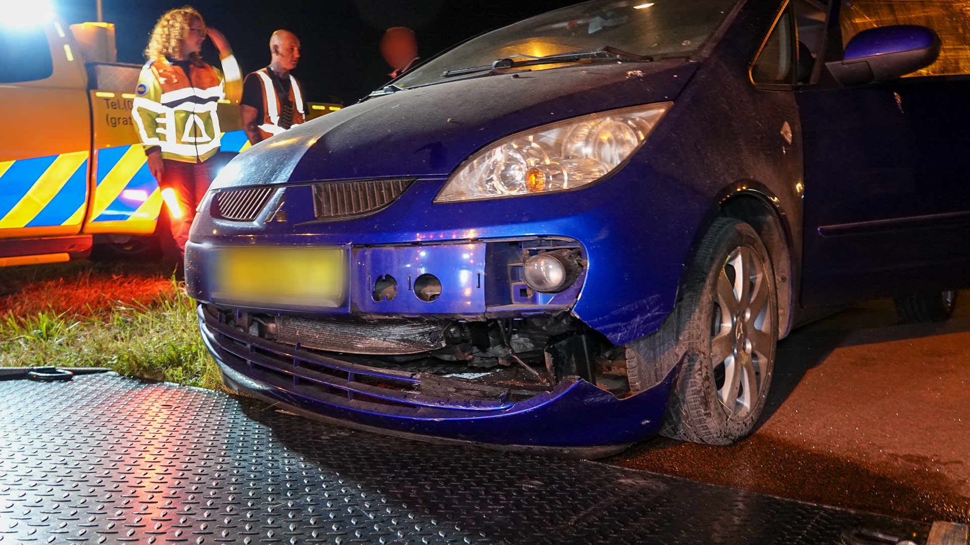 Een blauwe auto met zichtbare schade aan de bumper staat op een weg, met mensen in veiligheidsvesten en een hulpdienstvoertuig op de achtergrond in de nacht.