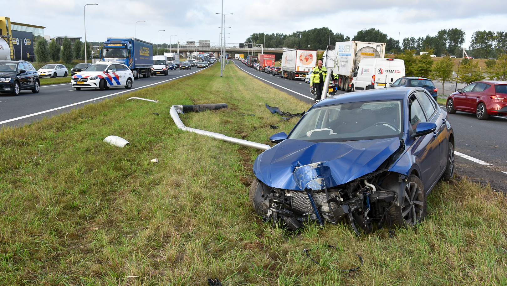 Beschadigde blauwe auto op een grasberm naast een snelweg, met een omvergereden lichtmast en politievoertuig in de achtergrond.