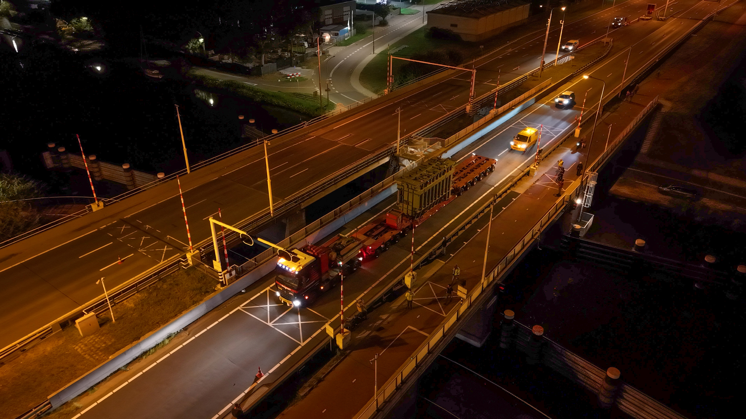 Luchtfoto van een nachtelijke weg met een grote vrachtwagen die een zware lading vervoert op een afgesloten rijstrook, omgeven door verkeersborden en verlichting.