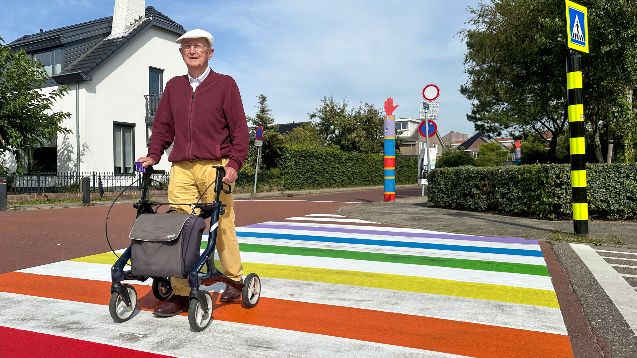 Een oudere man met een rollator steekt een zebrapad in regenboogkleuren over op een zonnige dag, met een huis en enkele verkeersborden op de achtergrond.