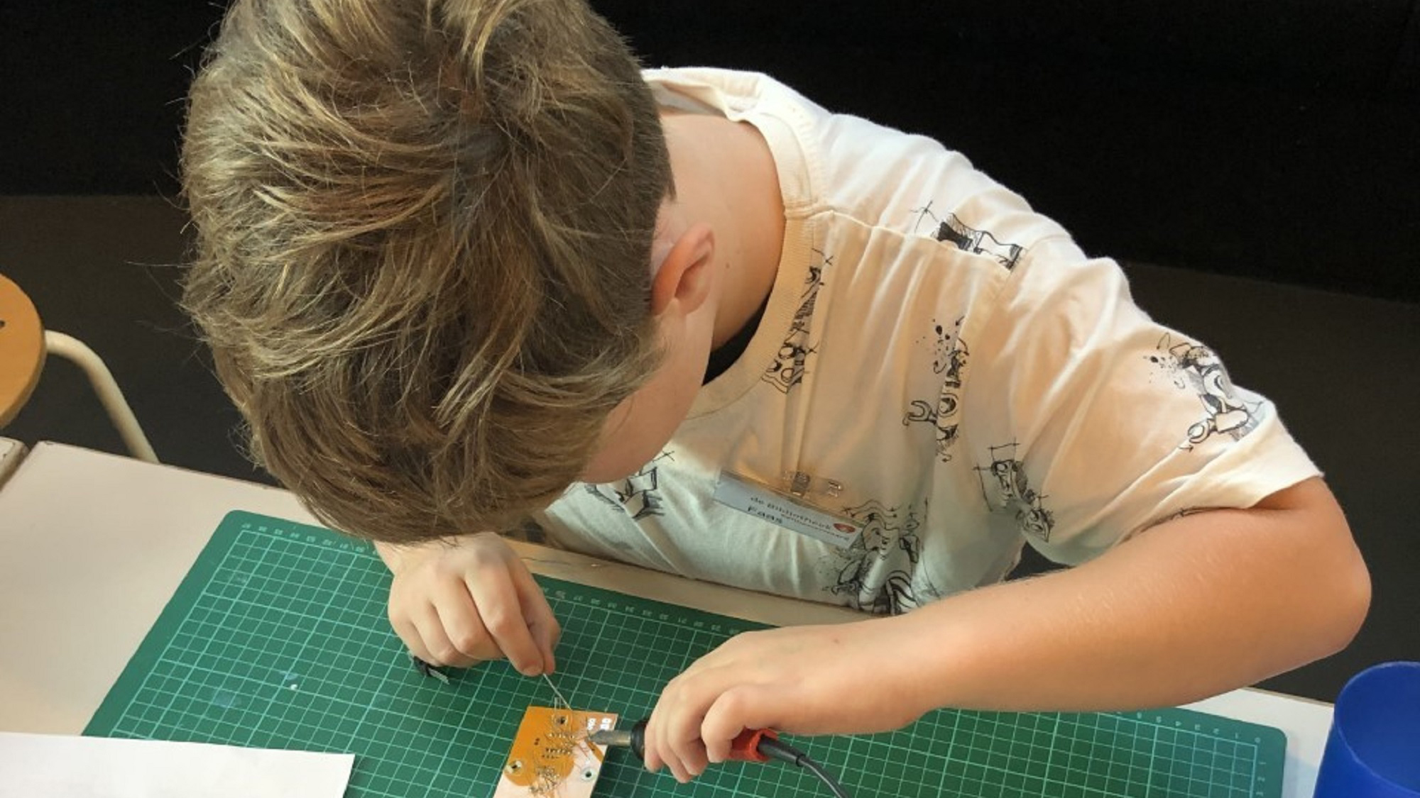Jonge jongen bezig met solderen van een elektronisch bord op een groene snijmat.