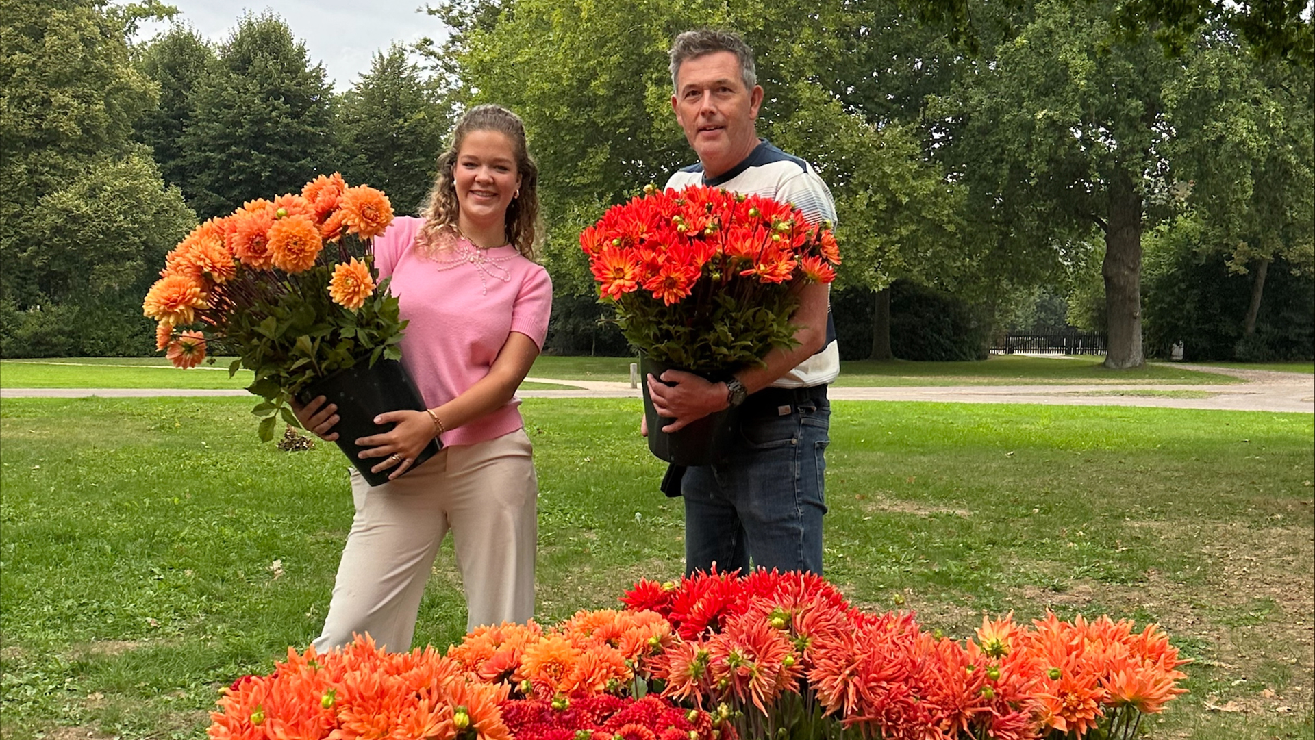 Twee mensen houden grote potten met oranje bloemen vast in een parkachtige omgeving, omringd door bomen en gras.