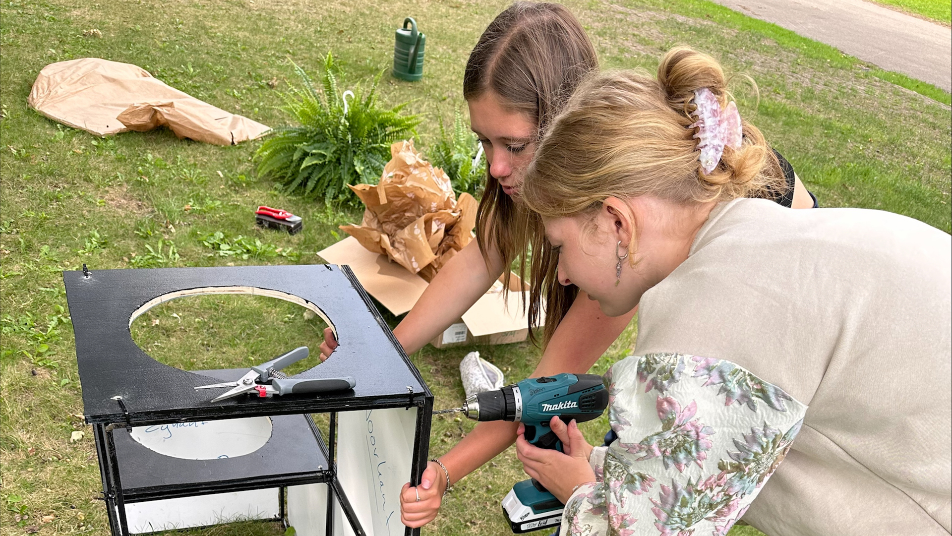 Twee meisjes werken aan een doe-het-zelf project op gras, één gebruikt een boormachine terwijl de ander met een schaartje werkt aan een zwart houten frame met ronde uitsnede.