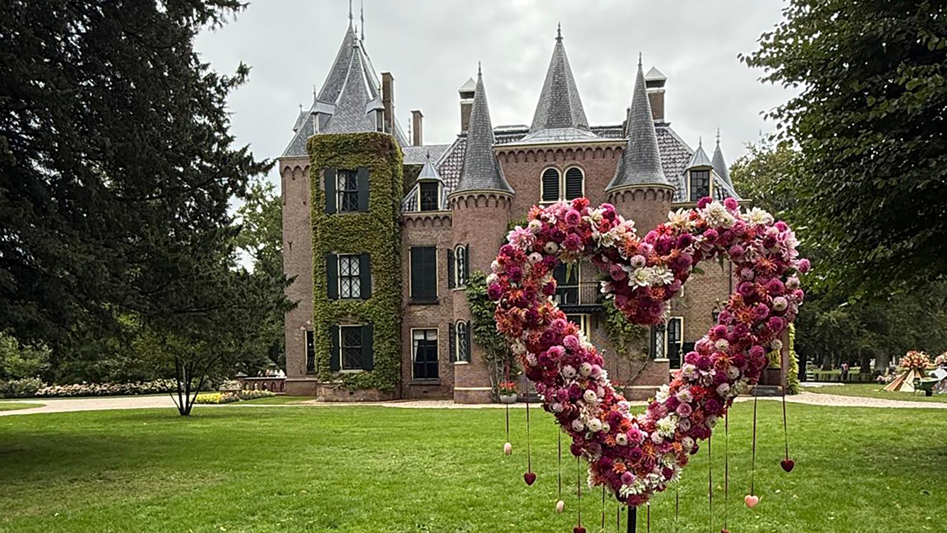 Een middeleeuws kasteel met ronde torens en een met klimop bedekte muur, op de voorgrond staat een groot hartvormig bloemenarrangement met roze en witte bloemen op een grasveld.