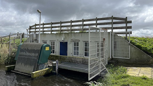 Een gemaal met metalen rooster en waterinlaat aan een kanaal, afgebeeld tegen een bewolkte hemel.