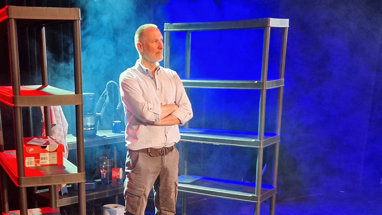 Een man met grijzend haar staat met zijn armen over elkaar naast lege metalen rekken op een podium, verlicht door blauw en rood licht.