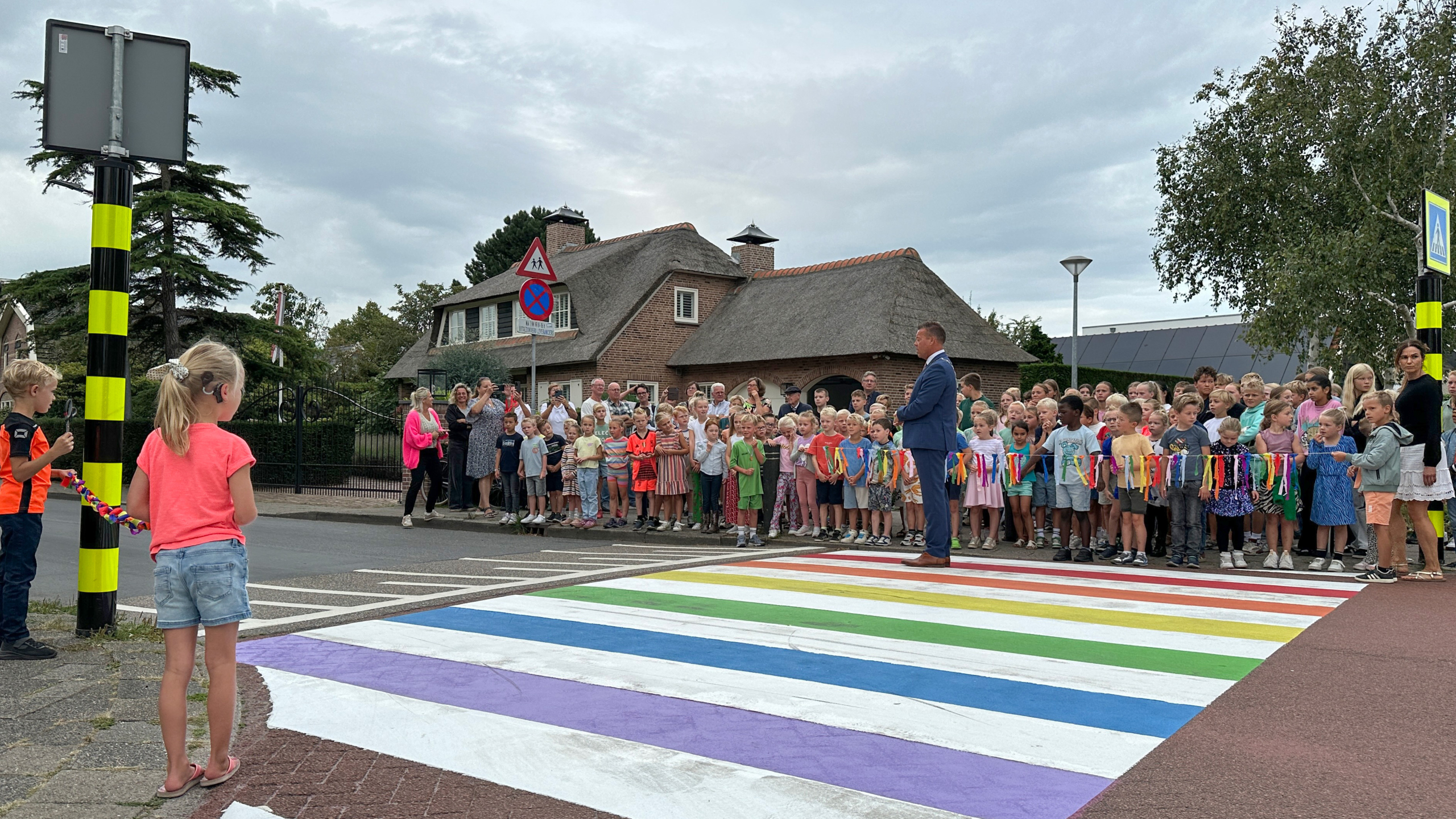 Kinderen en volwassenen verzamelen zich bij een regenboogzebrapad in een woonwijk, terwijl een man in een pak spreekt. Een meisje en jongen houden een lint vast om het pad officieel te openen.
