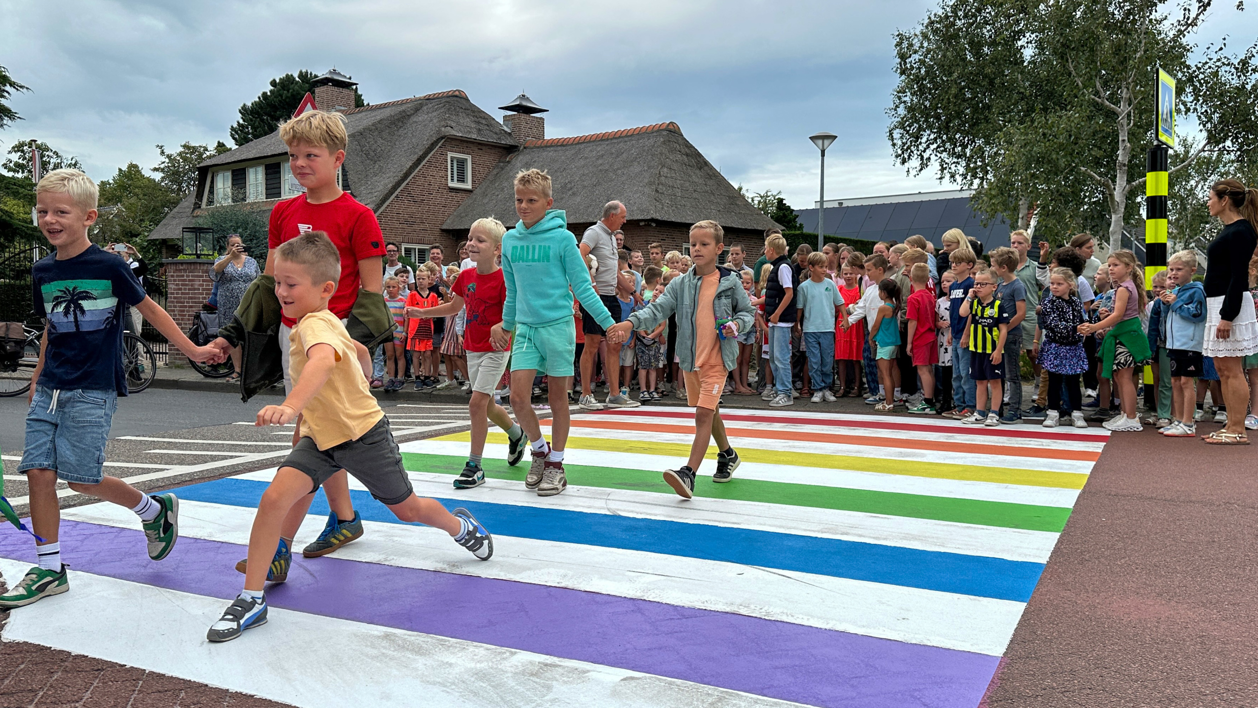 Een groep kinderen steekt een regenboogzebrapad over, gevolgd door een menigte mensen op de achtergrond.