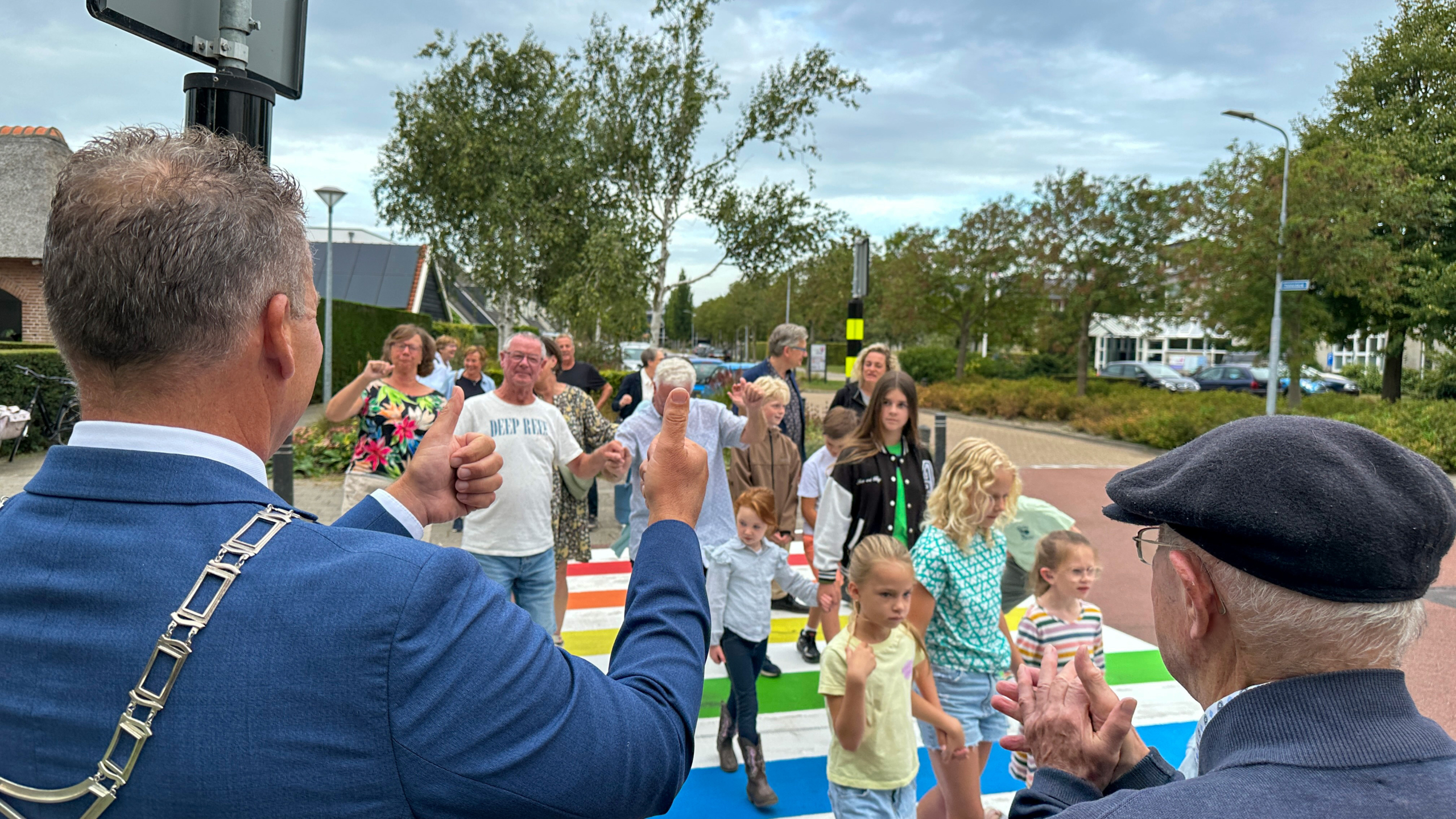 Mensen lopen over een kleurrijk zebrapad, begeleid door een man in een blauw pak die zijn duim omhoog steekt. Er zijn volwassenen en kinderen aanwezig, omgeven door een groenstedelijke omgeving met bomen en trottoirs.