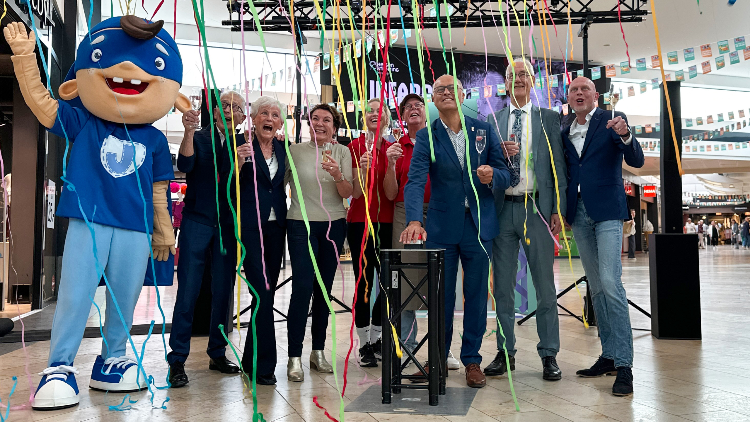 Een groep mensen viert een gebeurtenis in een winkelcentrum, omringd door linten, inclusief een groot maskerot in een blauw kostuum naast hen.