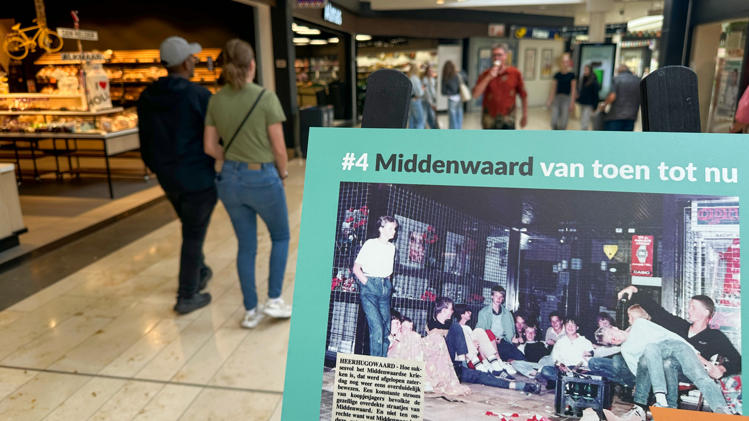 Twee personen lopen door een winkelcentrum; op de voorgrond is een bord te zien met een historische foto en de tekst 'Middenwaard van toen tot nu.'