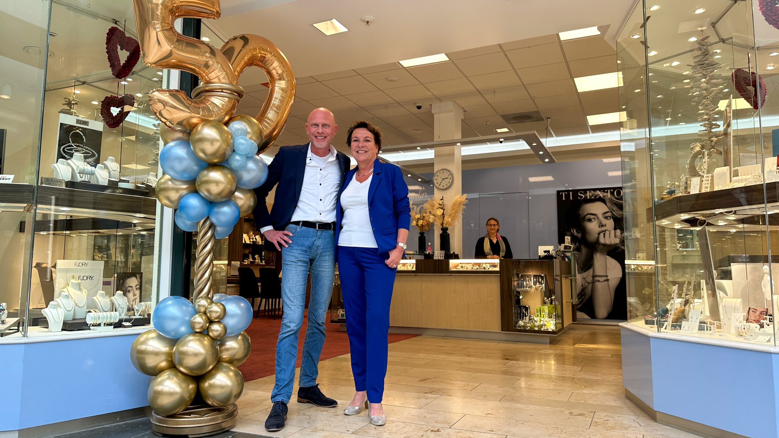 Twee mensen staan voor een juwelierszaak naast een grote ballonstal met het nummer 50, waarbij de winkel vitrines met sieraden toont.