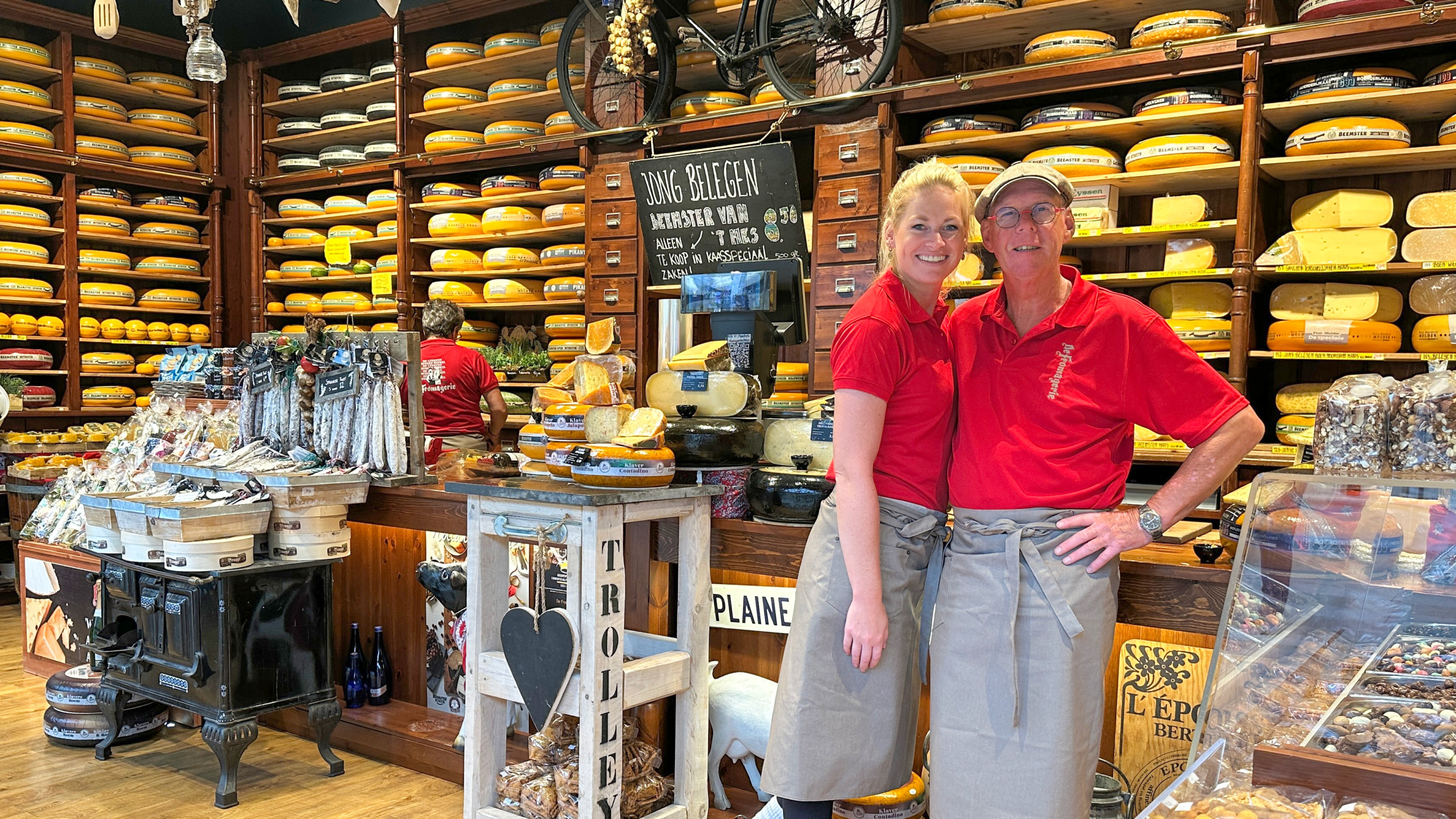 Twee medewerkers in een kaaswinkel, gekleed in rode shirts en grijze schorten, poseren voor planken vol ronde kazen.