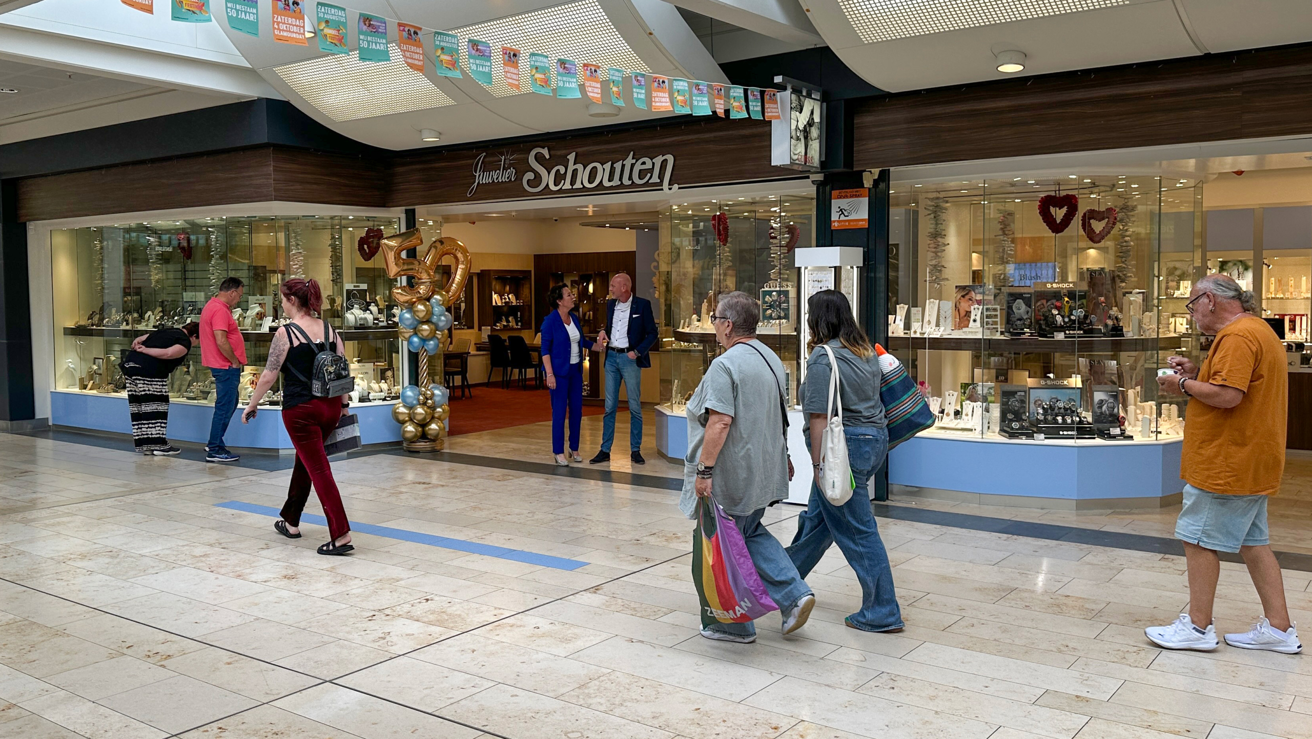 Winkelcentrum met een juwelierszaak genaamd Schouten, versierd met ballonnen en een getal 50, terwijl verschillende mensen voorbijlopen en het etalage bekijken.