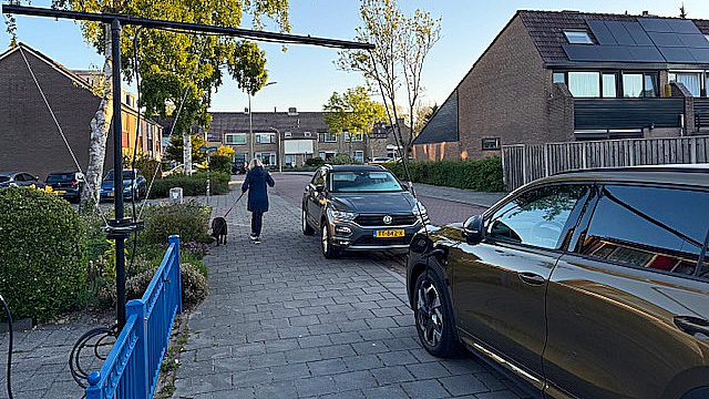 Straatbeeld met een auto geparkeerd op de stoep en een persoon die een hond uitlaat langs een pad, omringd door huizen met zonnepanelen en bomen.
