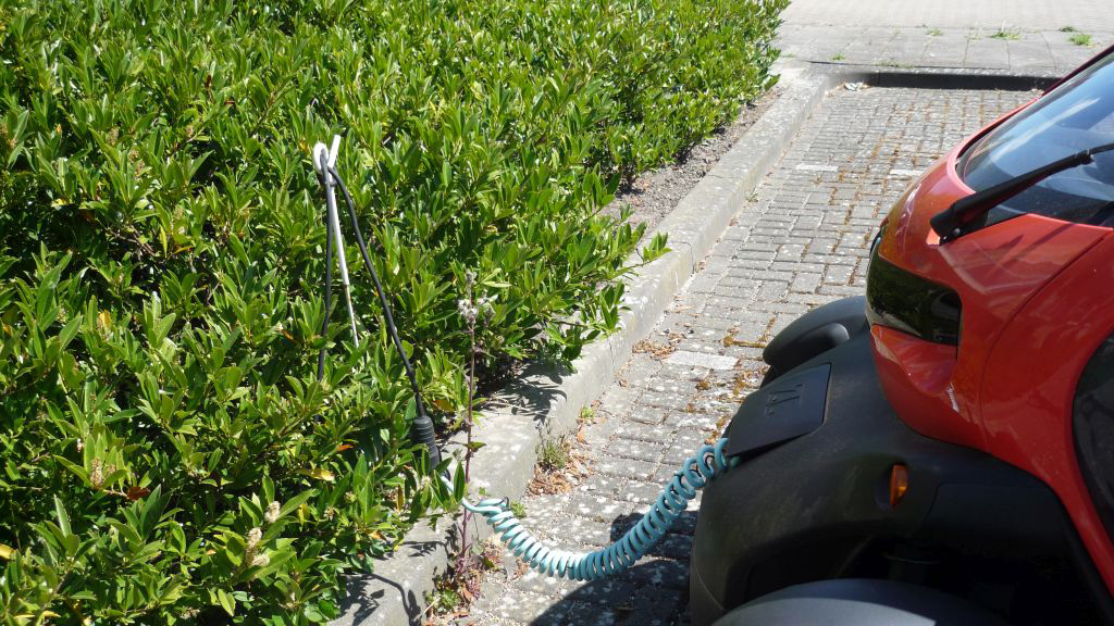 Een rode elektrische auto die staat geparkeerd op een parkeerplaats, is verbonden met een oplaadkabel die uit de nabije heg komt.