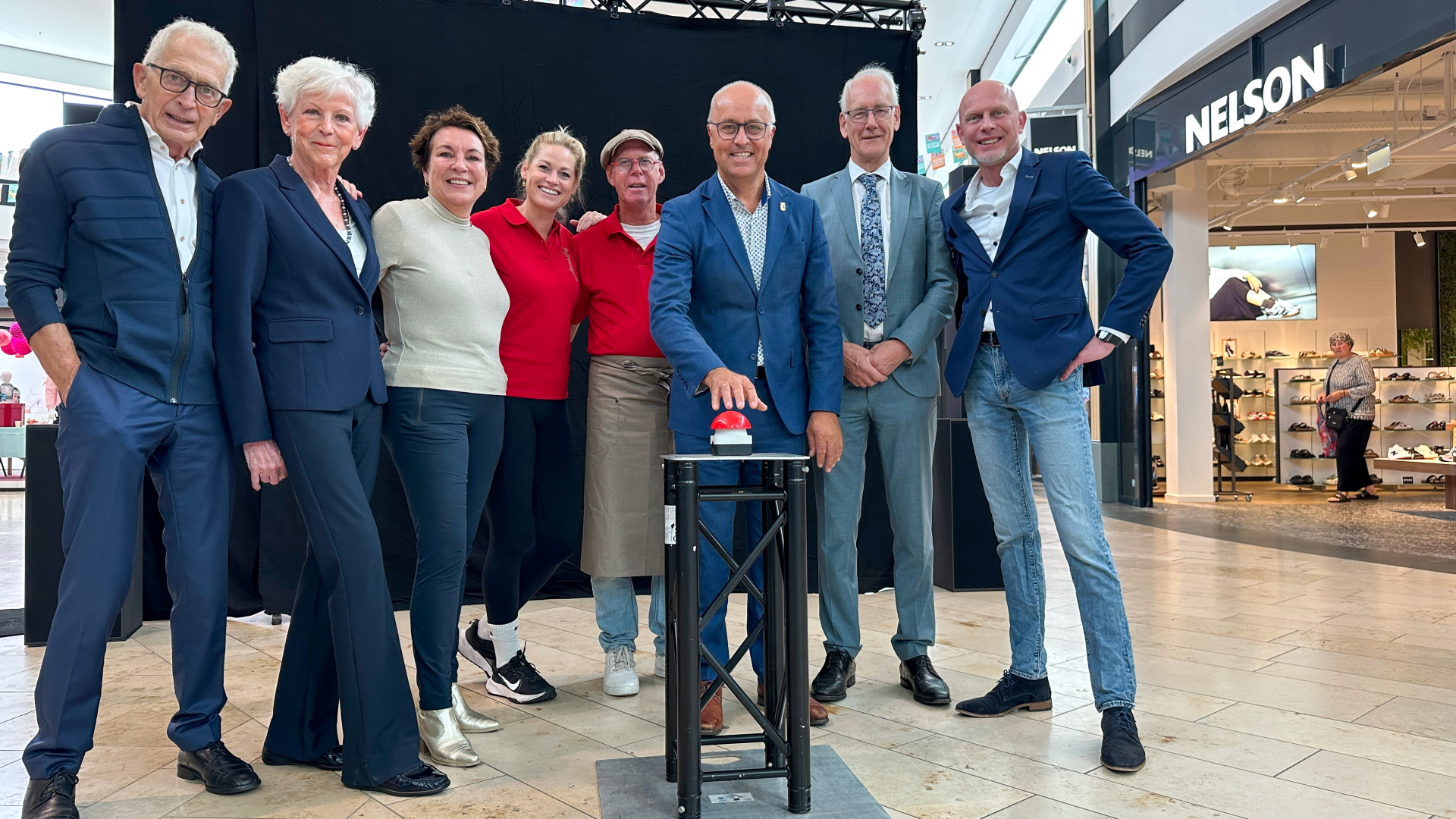 Een groep mensen poseert samen in een winkelcentrum naast een rode knop op een standaard, klaar om de knop in te drukken. Op de achtergrond is een Nelson-winkel zichtbaar.