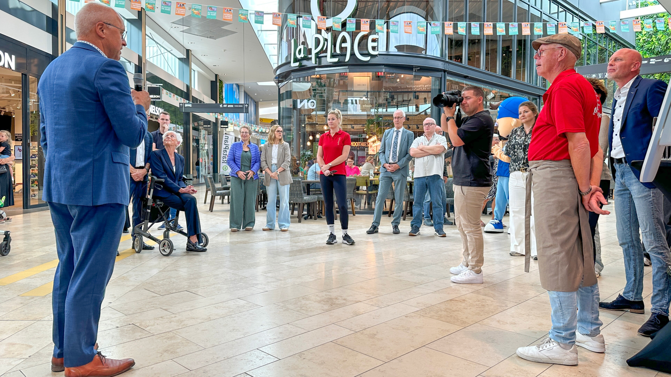 Een groep mensen verzameld in een winkelcentrum, terwijl een man in een blauw pak met een microfoon een toespraak houdt. In de achtergrond is een La Place café te zien. Mensen luisteren aandachtig, sommigen staan, anderen zitten. Een fotograaf maakt foto's van het tafereel.