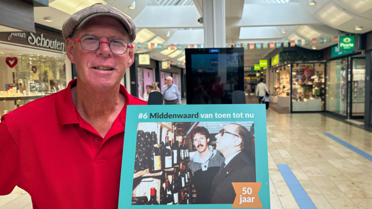 Een man in een rode polo en een grijze pet staat in een winkelcentrum en houdt een bord vast waarop de tekst 'Middenwaard van toen tot nu' staat, samen met een foto en tekst die verwijzen naar een 50-jarig jubileum.