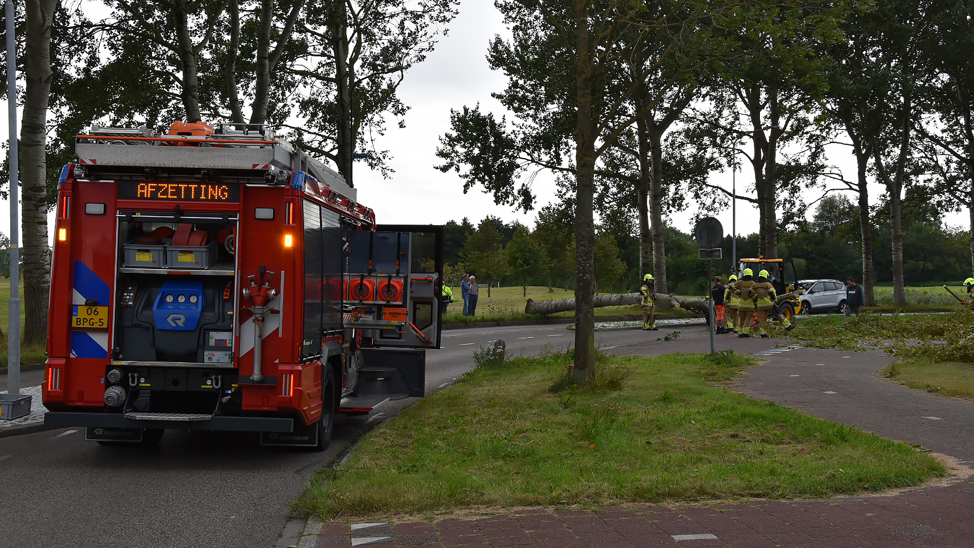 Brandweerauto met het woord 'AFZETTING' op de display, geparkeerd langs een weg waar een omgevallen boom ligt en brandweerlieden bezig zijn met opruimen.
