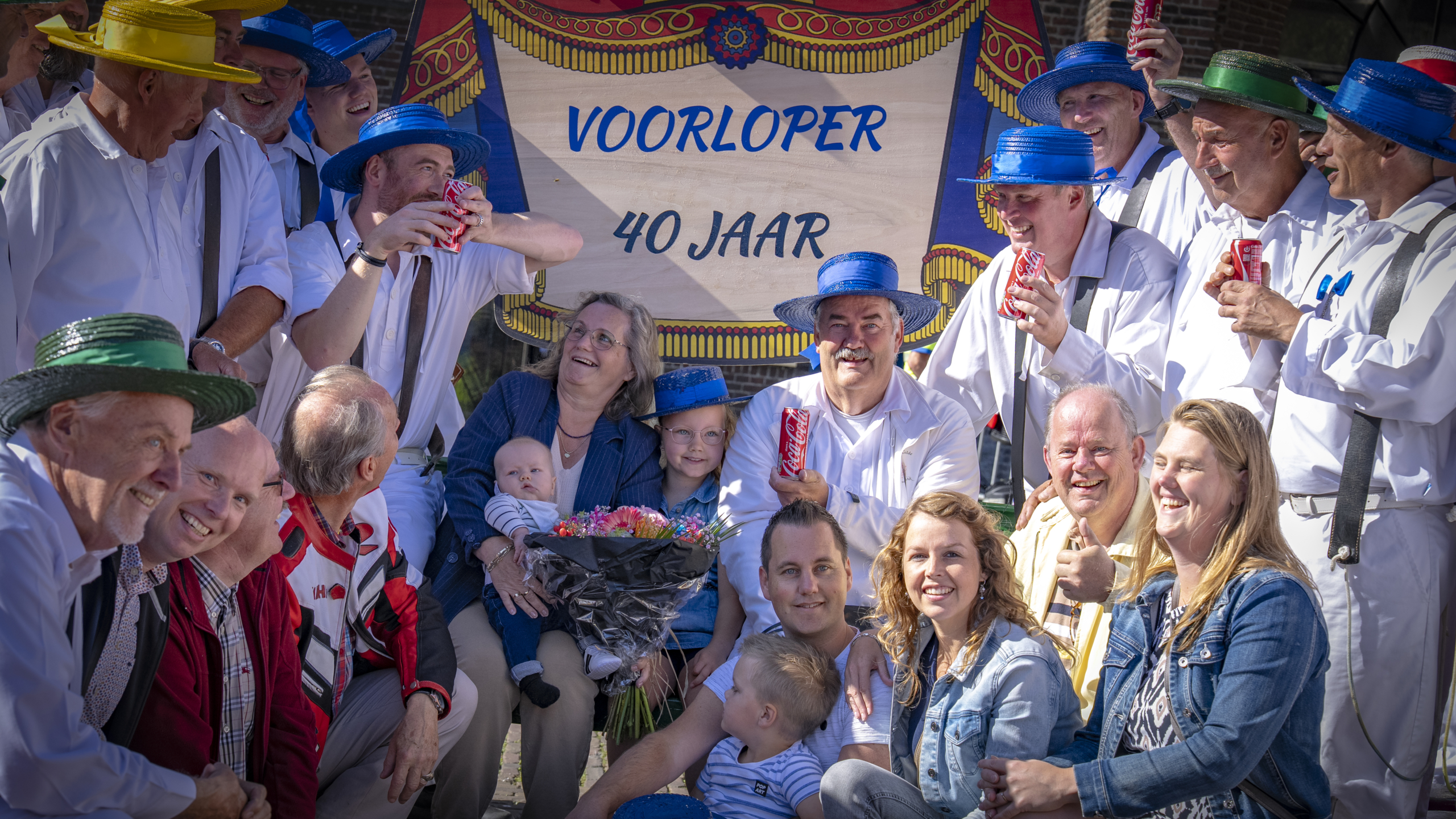 Groep mensen poseert vrolijk voor een spandoek met de tekst 'VOORLOPER 40 JAAR', waarbij sommigen kleurrijke hoeden dragen en blikjes frisdrank vasthouden; een vrouw houdt een baby en een boeket bloemen vast, omringd door lachende volwassenen en kinderen.