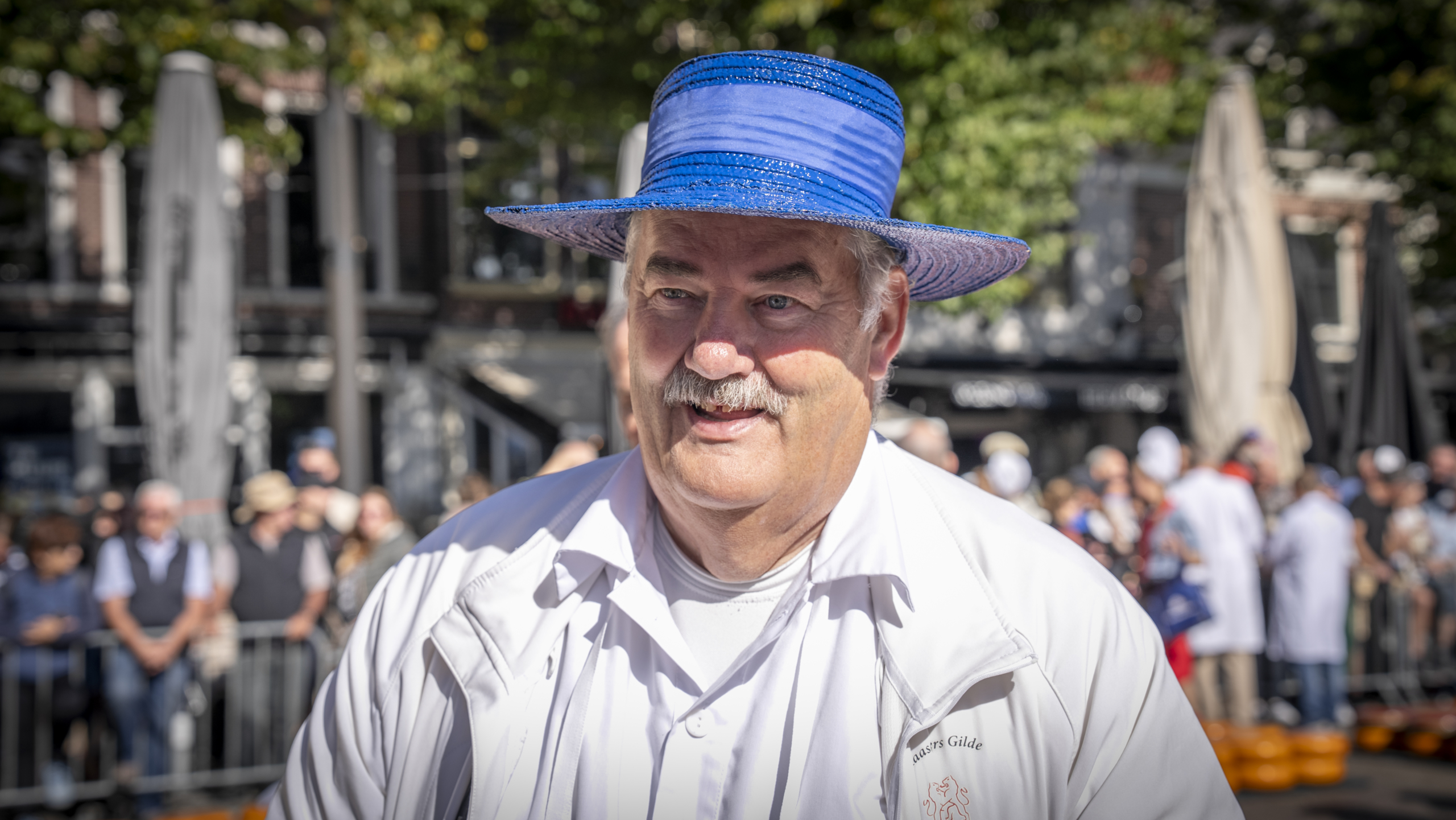 Een man met een blauwe hoed en witte jas tijdens een buitenactiviteit, met een menigte op de achtergrond.