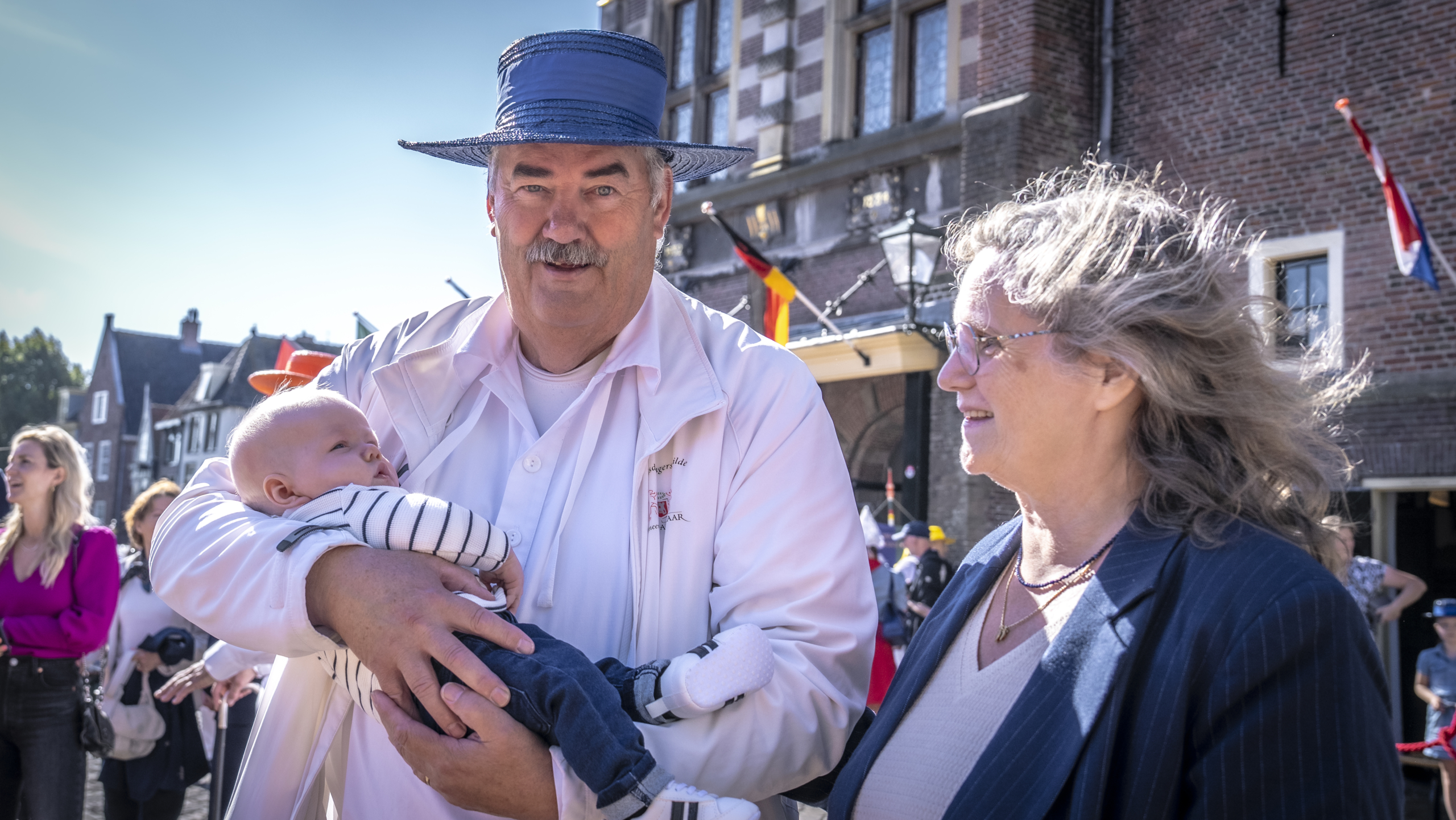 Een man in een witte jas en blauwe hoed houdt een baby in zijn armen, terwijl een vrouw met bril hem glimlachend aankijkt op straat, met historische gebouwen op de achtergrond.