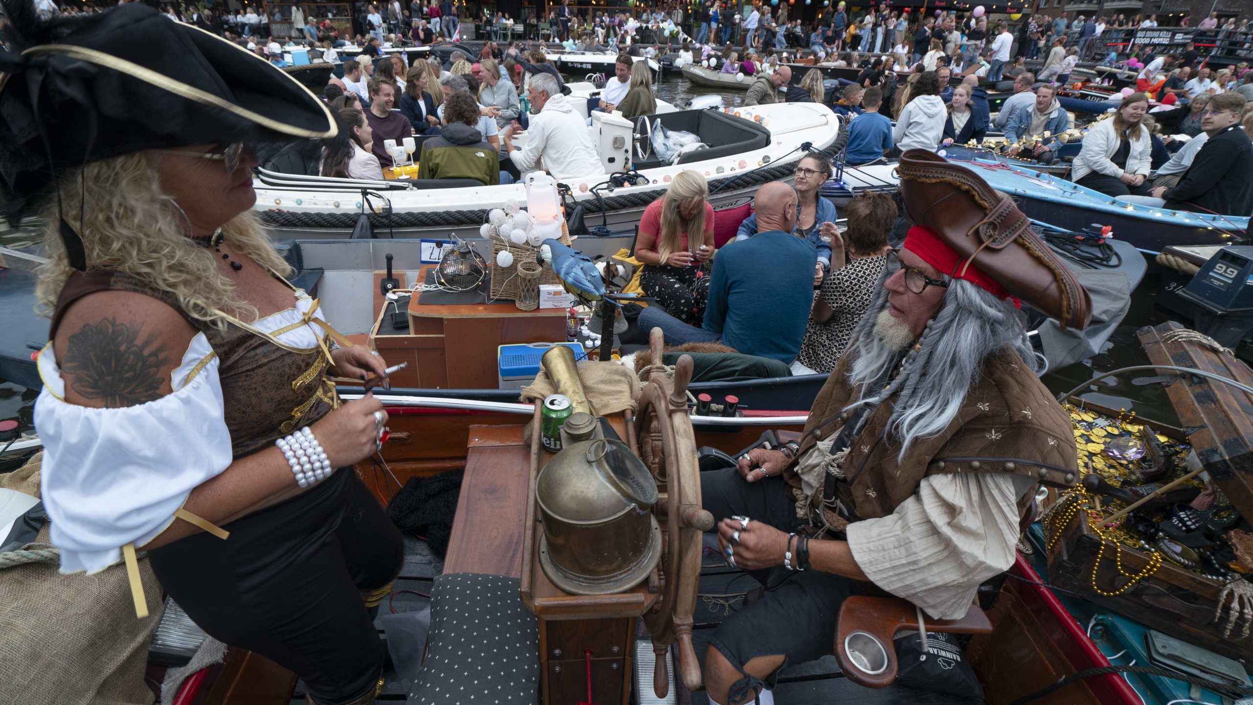 Twee mensen verkleed als piraten zitten op een drukke boot tijdens een festival. De vrouw draagt een zwarte hoed en bruine corset, terwijl de man een rode hoofdband en piratenhoed draagt. Om hen heen zitten mensen op andere boten, terwijl op de achtergrond een drukte van mensen zichtbaar is. Er staat een stuurwiel op de boot en een papegaai zit op de reling.