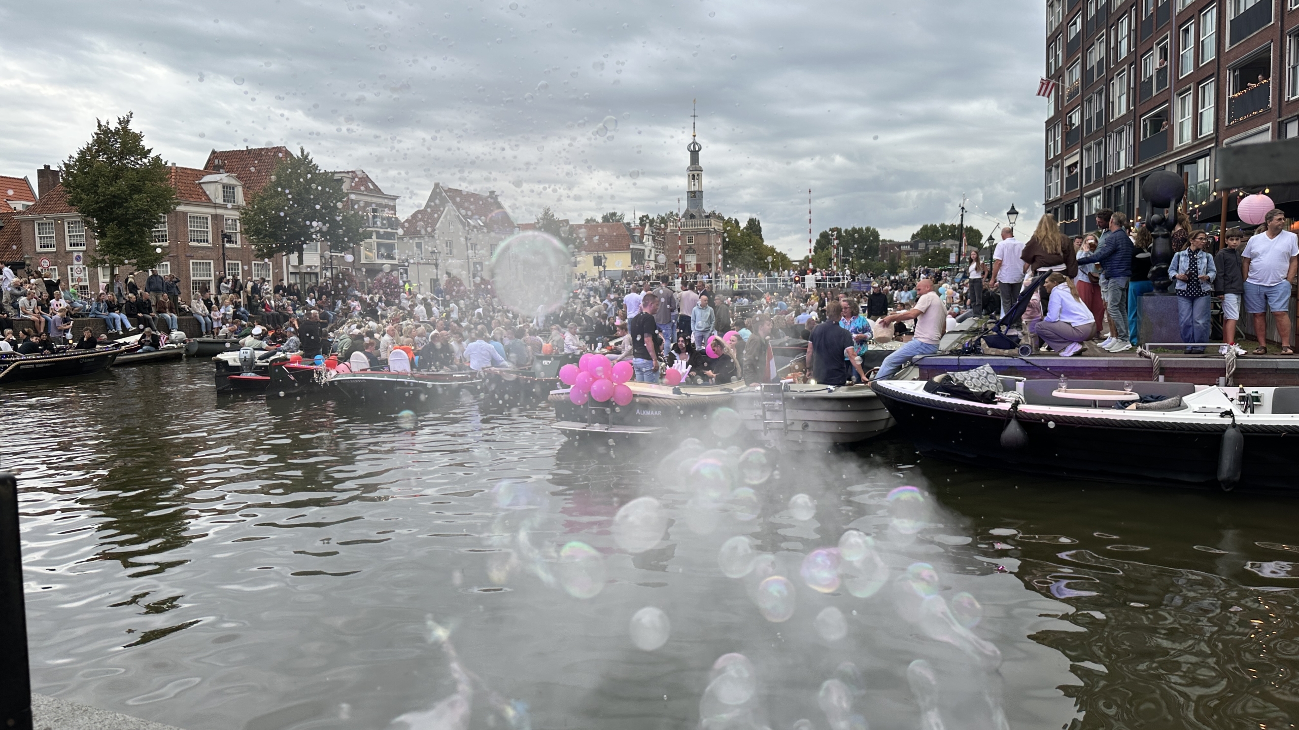 Drukke scène op een grachtenfestival met mensen in boten, omringd door toeschouwers op de kade. Er zweven veel bellen in de lucht en er zijn roze ballonnen op één van de boten. Op de achtergrond zijn historische gebouwen en een toren zichtbaar.