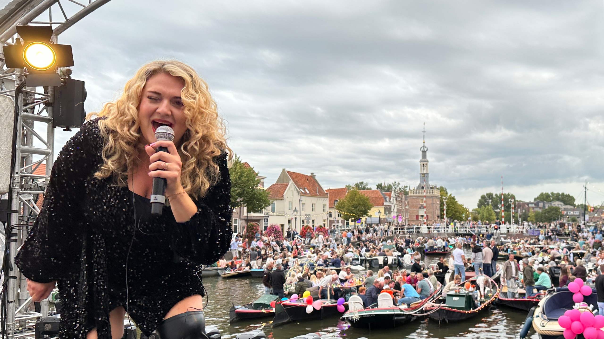 Een vrouw met krullend blond haar zingt in een microfoon op een podium, terwijl op de achtergrond een drukke bijeenkomst van boten en mensen plaatsvindt bij een waterweg in een stad.