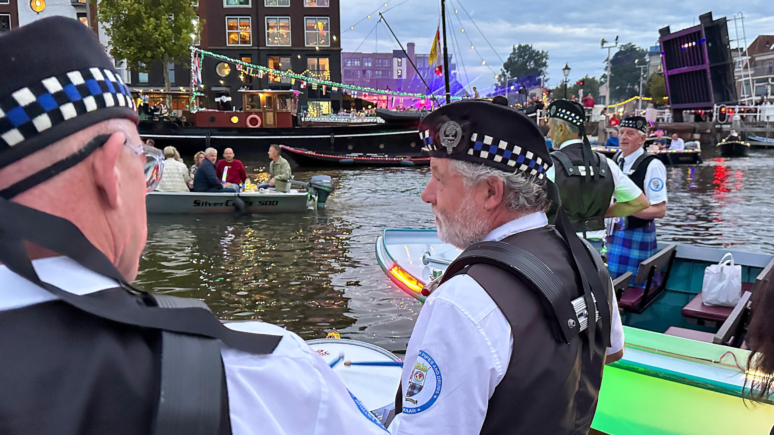 Mannen in traditionele Schotse uniformen op een boot in een sfeervol verlichte gracht, met mensen aan de oever en andere boten op de achtergrond.