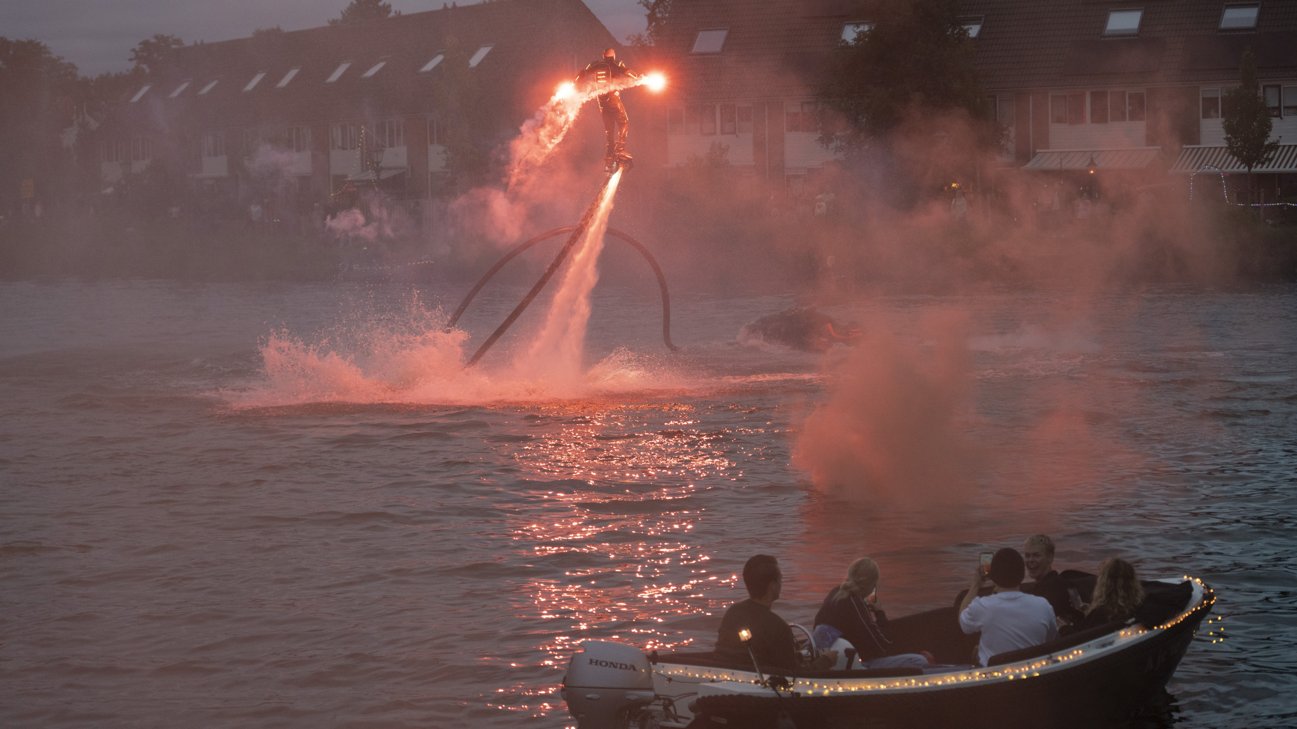 Een persoon staat op een flyboard boven het water, met rook en fakkels in beide handen, omringd door rood licht, terwijl een kleine boot met toeschouwers op de voorgrond te zien is.