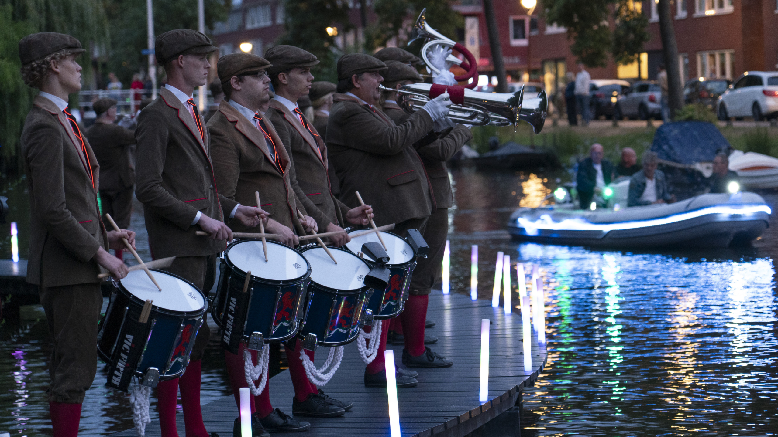 Een groep muzikanten in bruine pakken en petten speelt naast een gracht, met trommels en koperblaasinstrumenten. Op de achtergrond is een boot op het water te zien, versierd met lichtjes.